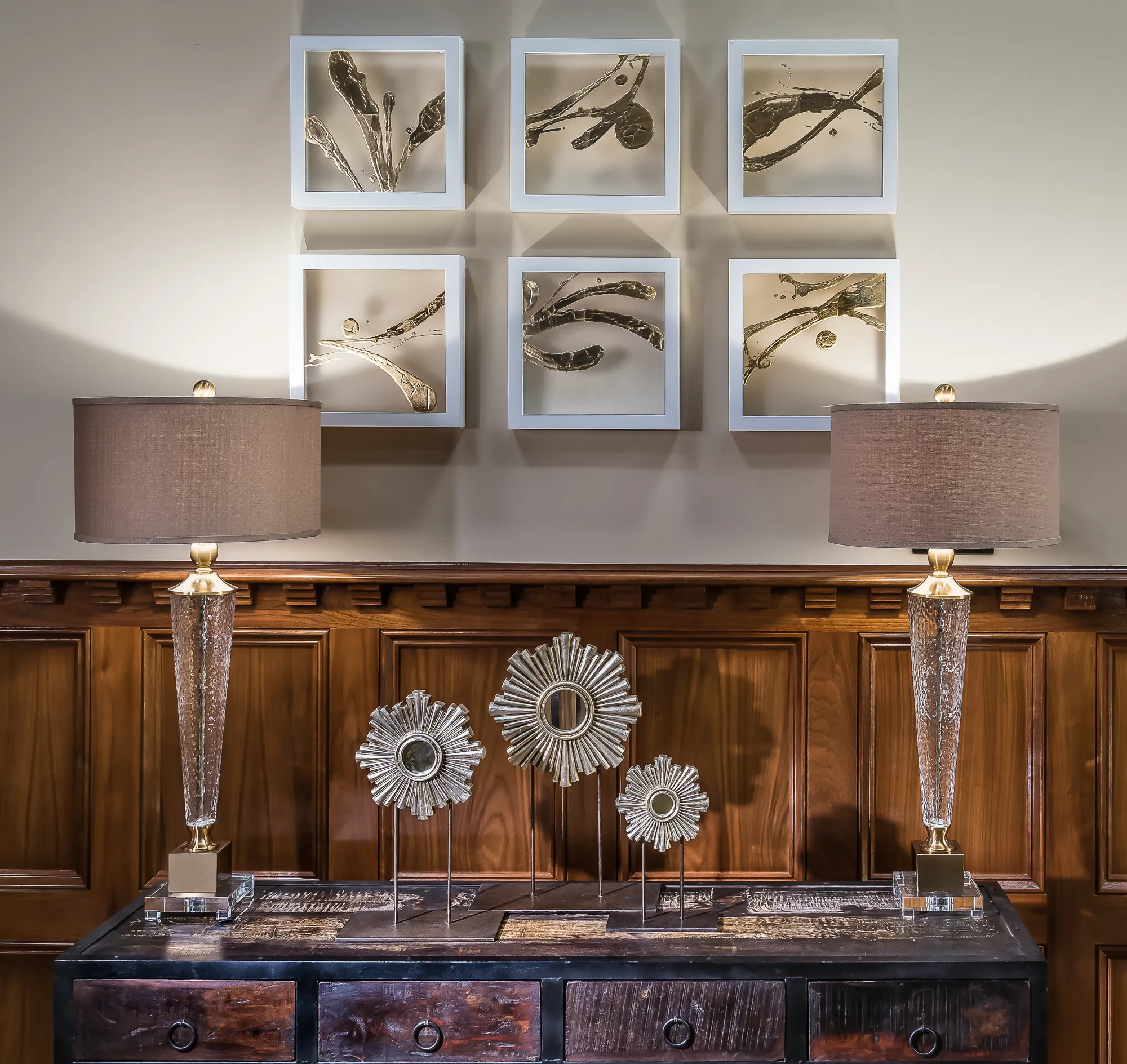 Two tall glass lamps and three metal sunburst sculptures on a console table are below six square frames displaying abstract gold and white wall art.