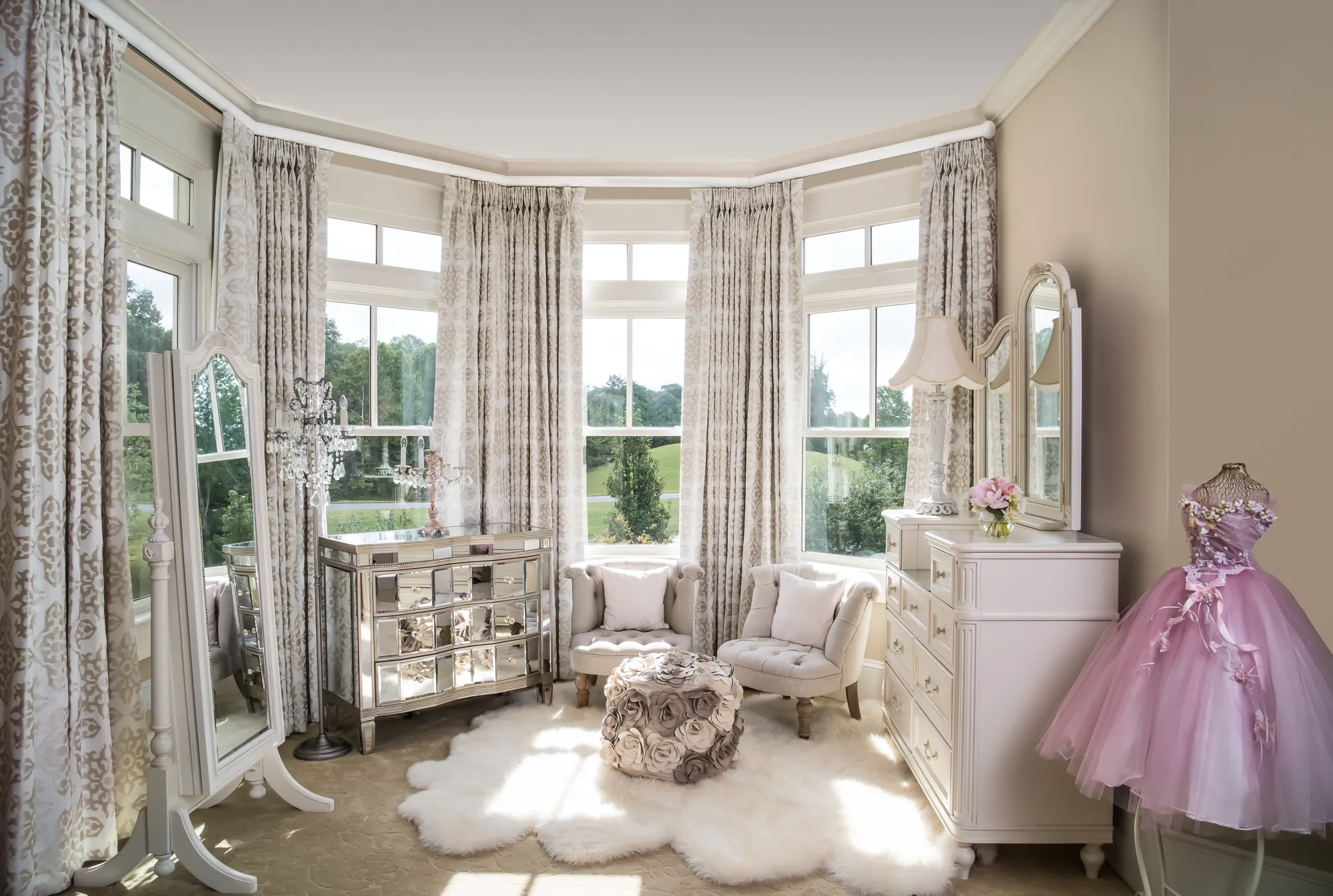 A luxurious dressing room with a large bay window, mirrored chest of drawers, two light gray tufted chairs, and a pink tulle gown displayed on a mannequin.