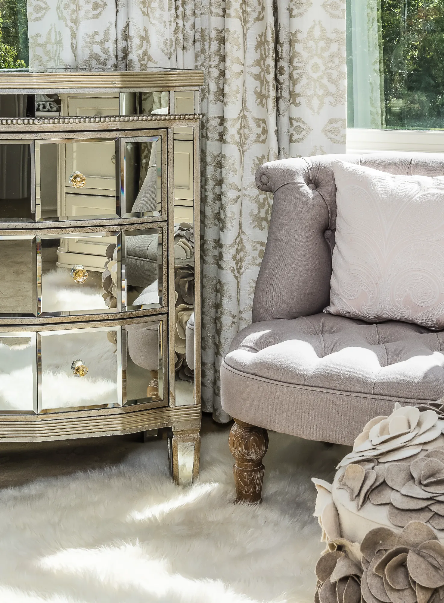 A close-up of a mirrored dresser and a tufted neutral chair, placed on a white faux fur rug next to light filtering through patterned curtains.