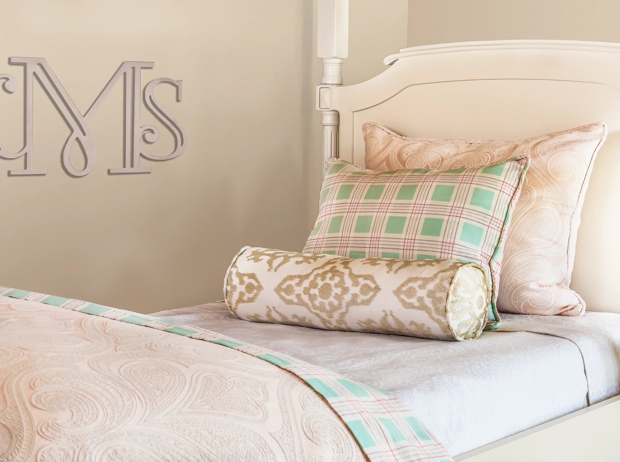 A white bed with pink paisley bedding and layered decorative pillows in plaid and brocade patterns. Large silver initials "MS" decorate the beige wall.