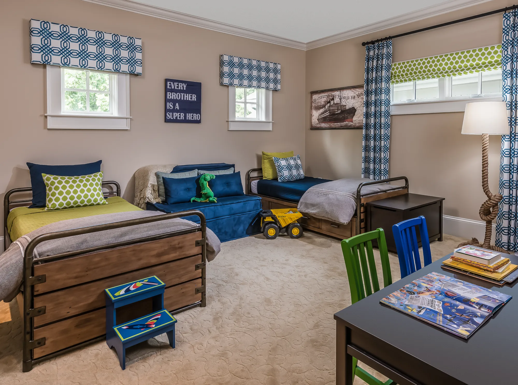 A shared children's bedroom features two rustic wood and metal beds and a navy daybed, all covered in blue and green bedding. A sign reads, "EVERY BROTHER IS A SUPER HERO." A small desk with bright chairs is in the foreground.
