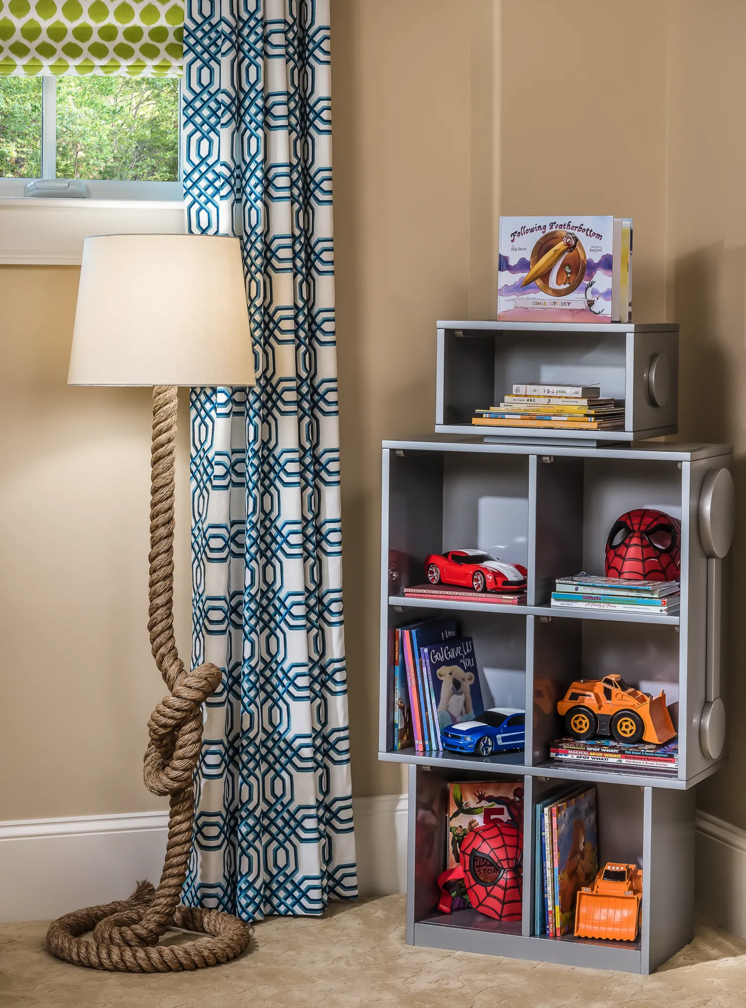 A bright, decorated child's room featuring a rope-base floor lamp and a gray cubby shelf holding colorful books, toy cars, and a Spiderman mask.