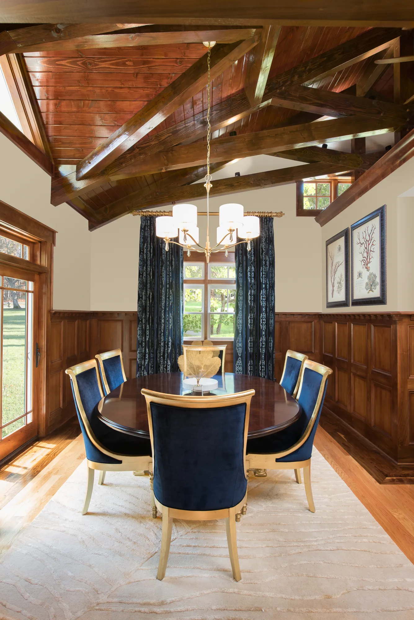 A formal dining room featuring dark wood wainscoting, exposed wooden ceiling beams, and a light area rug. A round dark table is set with six gold-framed chairs upholstered in rich blue velvet.