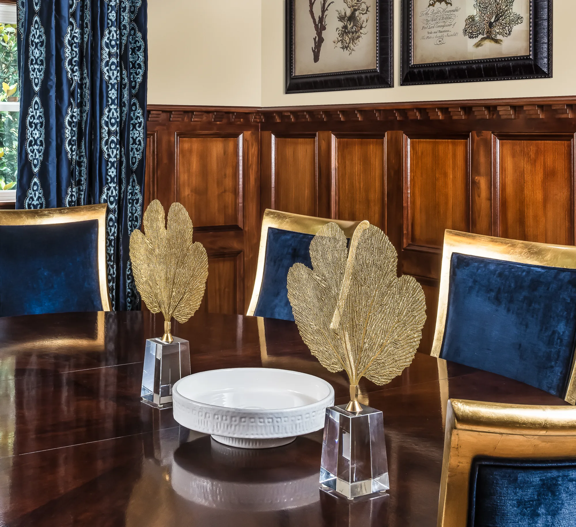 A rich, formal dining room with blue velvet chairs, dark wood paneling, and a polished table centered with gold coral sculptures.