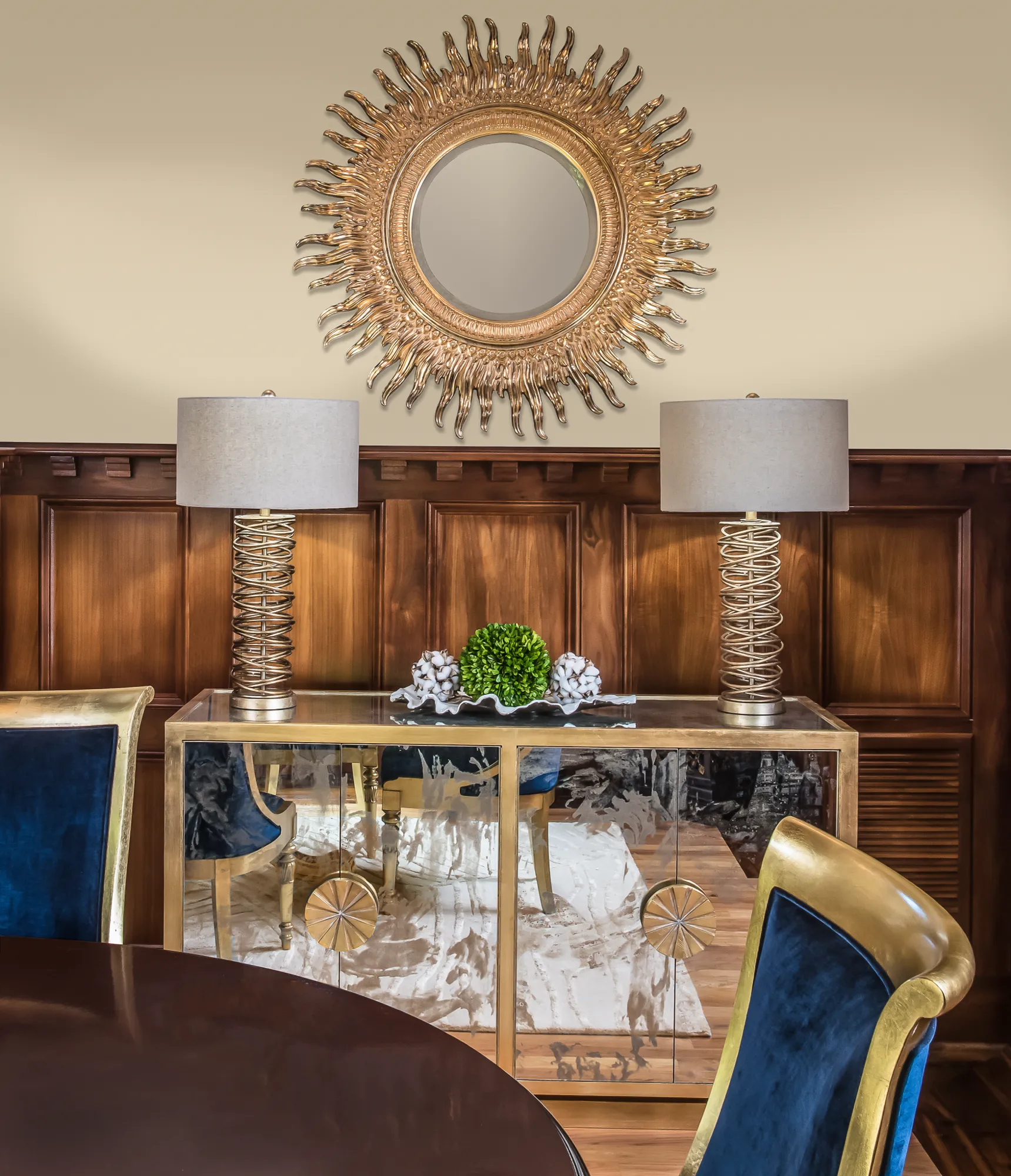 A sunburst mirror hangs above a gold-framed mirrored console flanked by coiled lamps, set against dark wood paneling and velvet dining chairs.