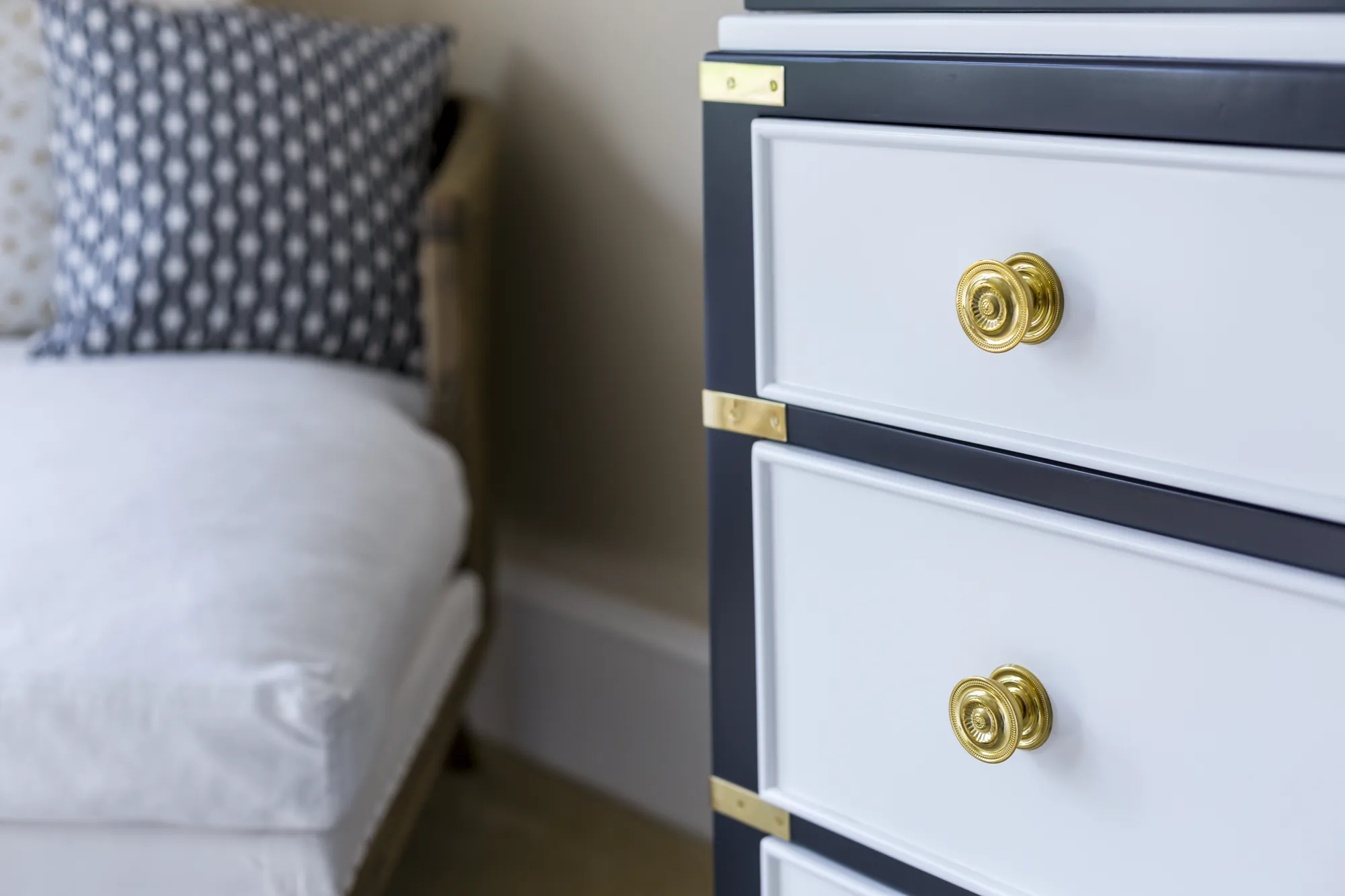 Close-up view of a navy and white dresser detailed with gold brass campaign hardware, next to a white chaise lounge and patterned pillow.