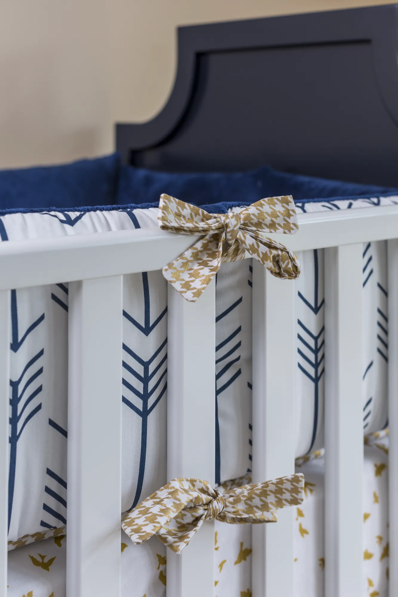 A white nursery crib featuring a navy headboard and blue arrow-patterned bedding tied with decorative gold and white bows.