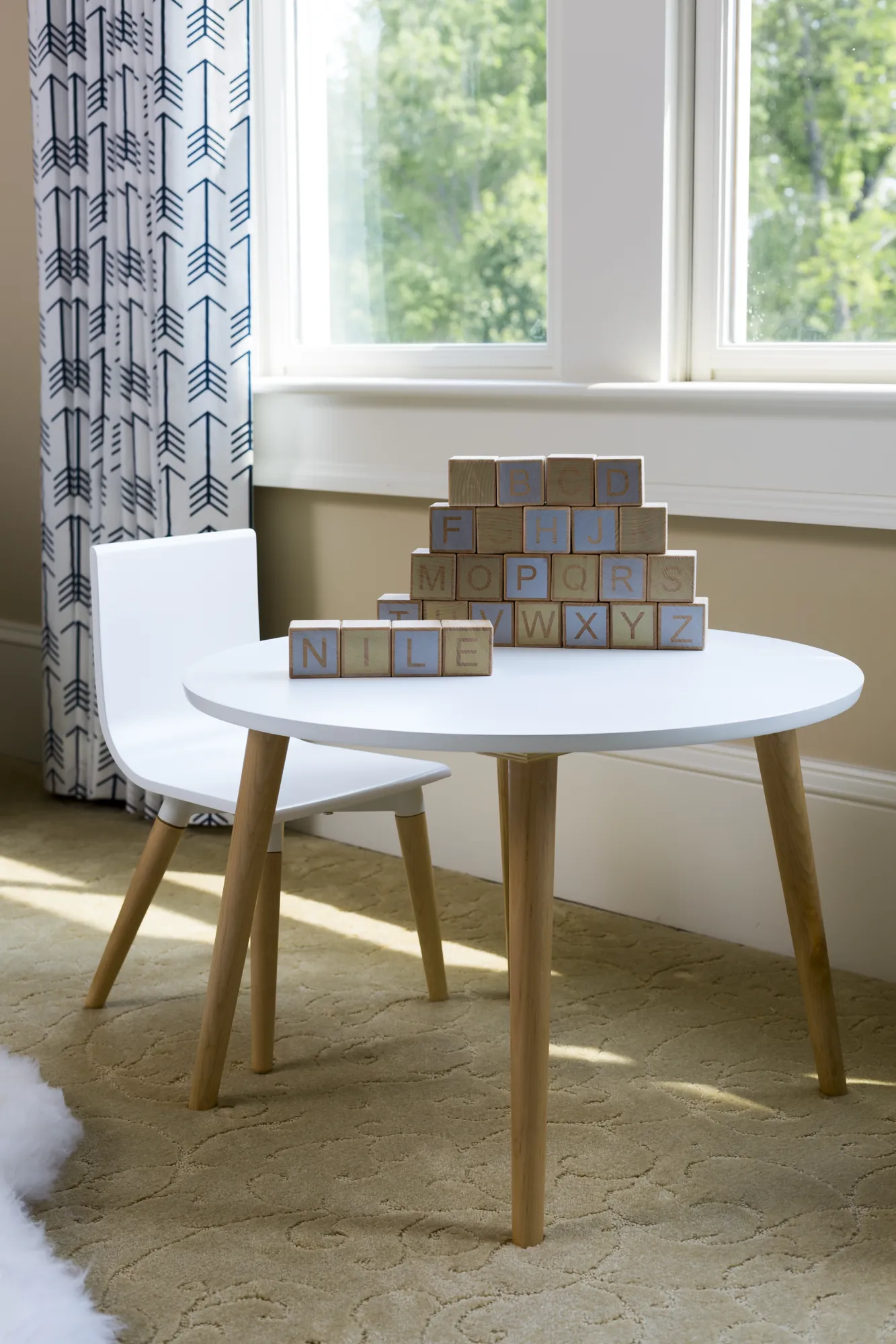 A child's white table and chair are in front of a window with white curtains printed with blue arrows. A pyramid of wooden alphabet blocks is stacked on the table.