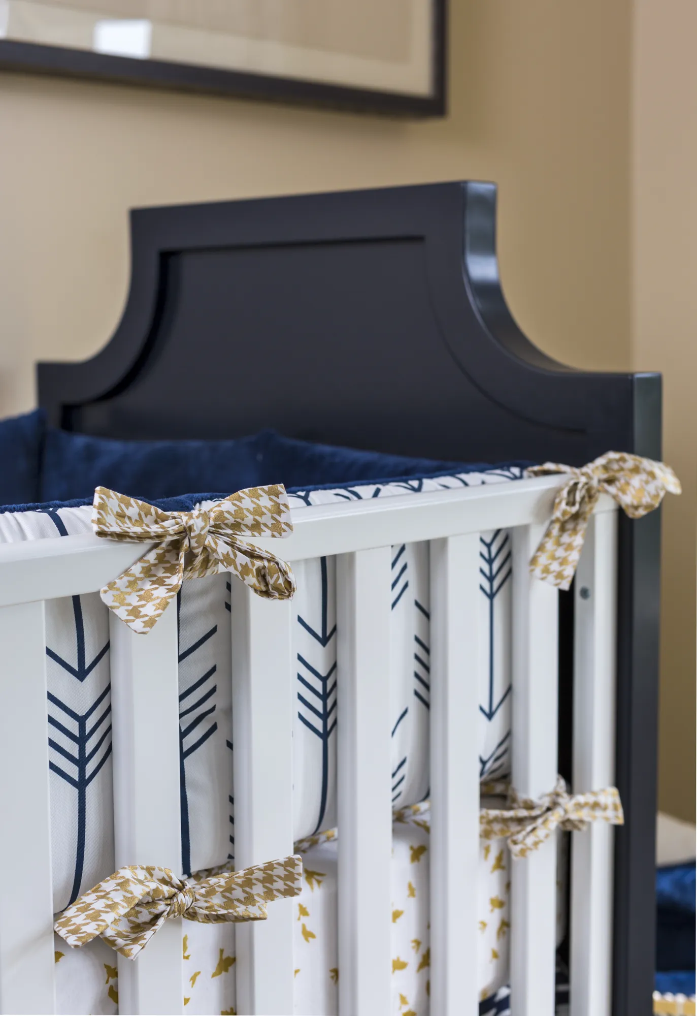 A close-up of a white crib with a navy headboard, featuring a bumper pad with a navy arrow pattern secured by gold-patterned bow ties.