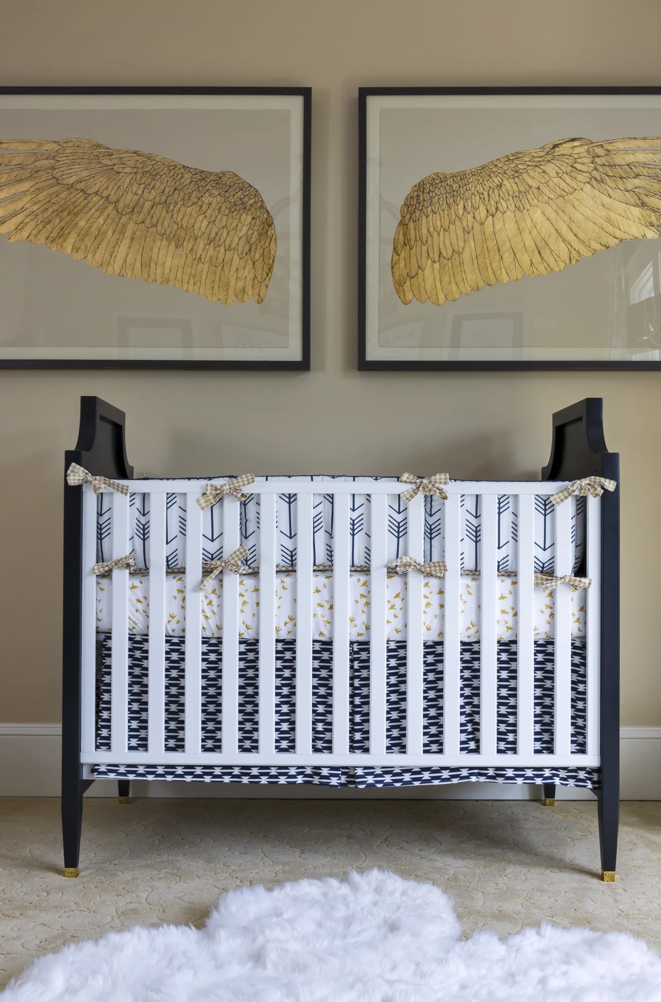 A navy and white crib with geometric bedding sits in a modern nursery. Above it, two framed gold wing prints hang on a tan wall over a fluffy white rug.