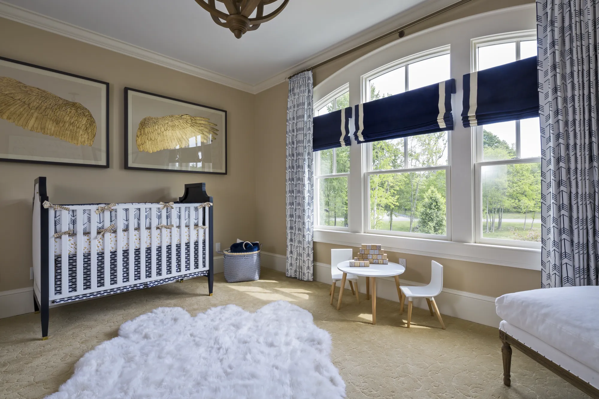 An elegant nursery featuring a black and white crib under two framed golden wings. A white fluffy rug covers the floor near a small table with toy blocks by large, bright arched windows.