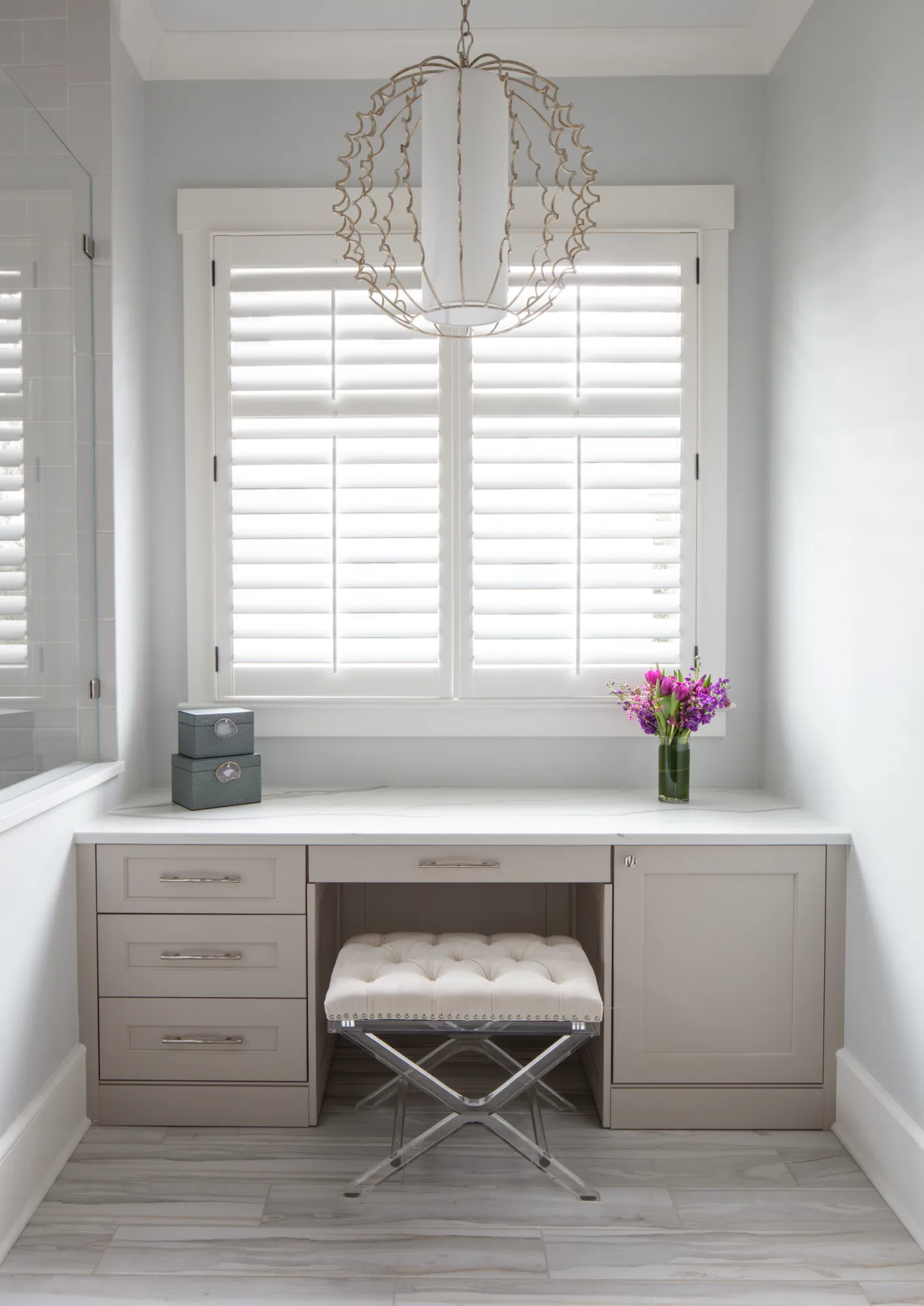 A bright, elegant dressing niche features light taupe built-in cabinets, a white countertop, and a cream tufted stool with a chrome base. An ornate chandelier hangs above the white shuttered window.