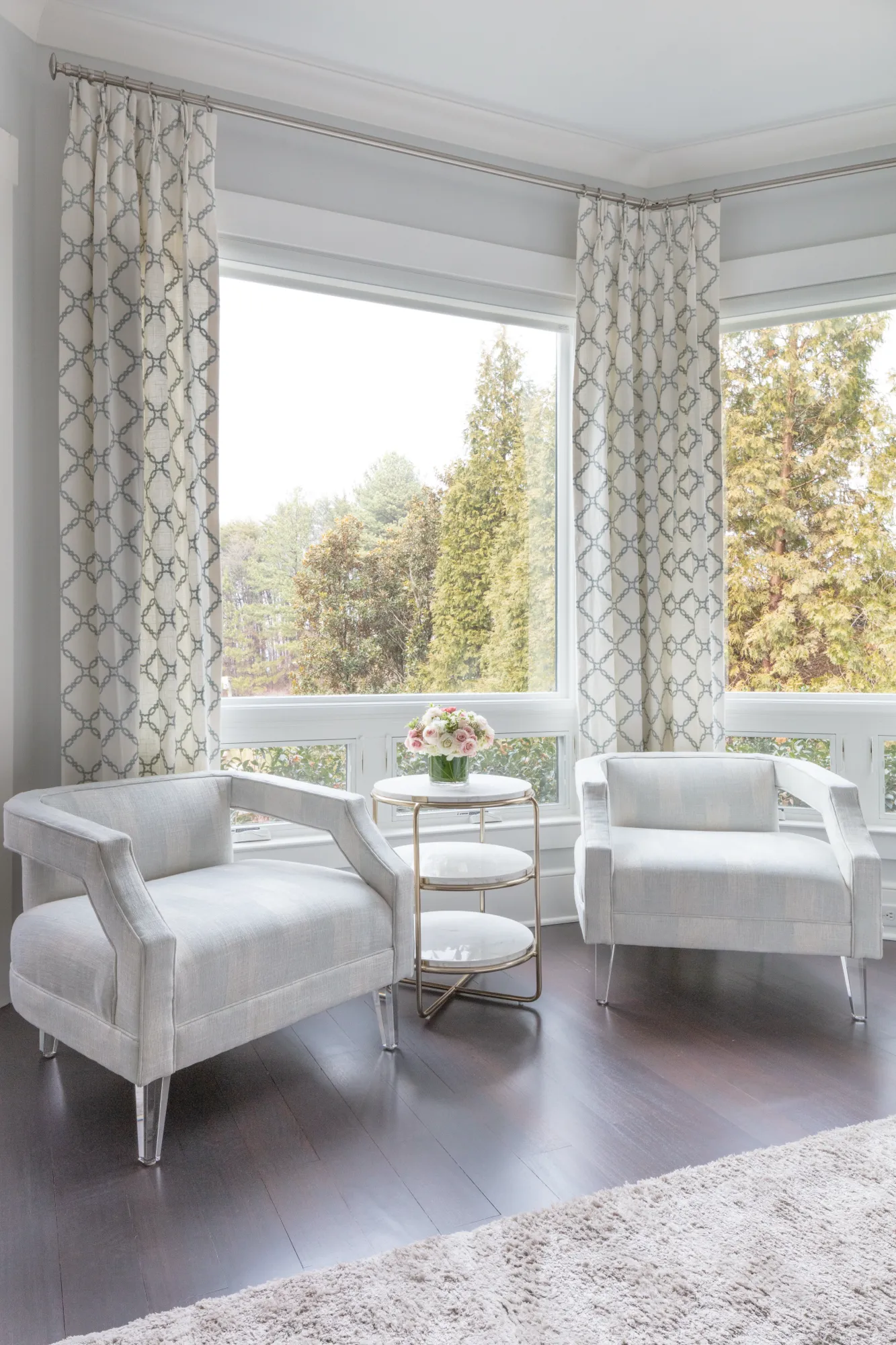 Two matching light gray accent chairs frame a small tiered marble side table with roses, set in a bright space with dark floors and large windows showing a wooded landscape.