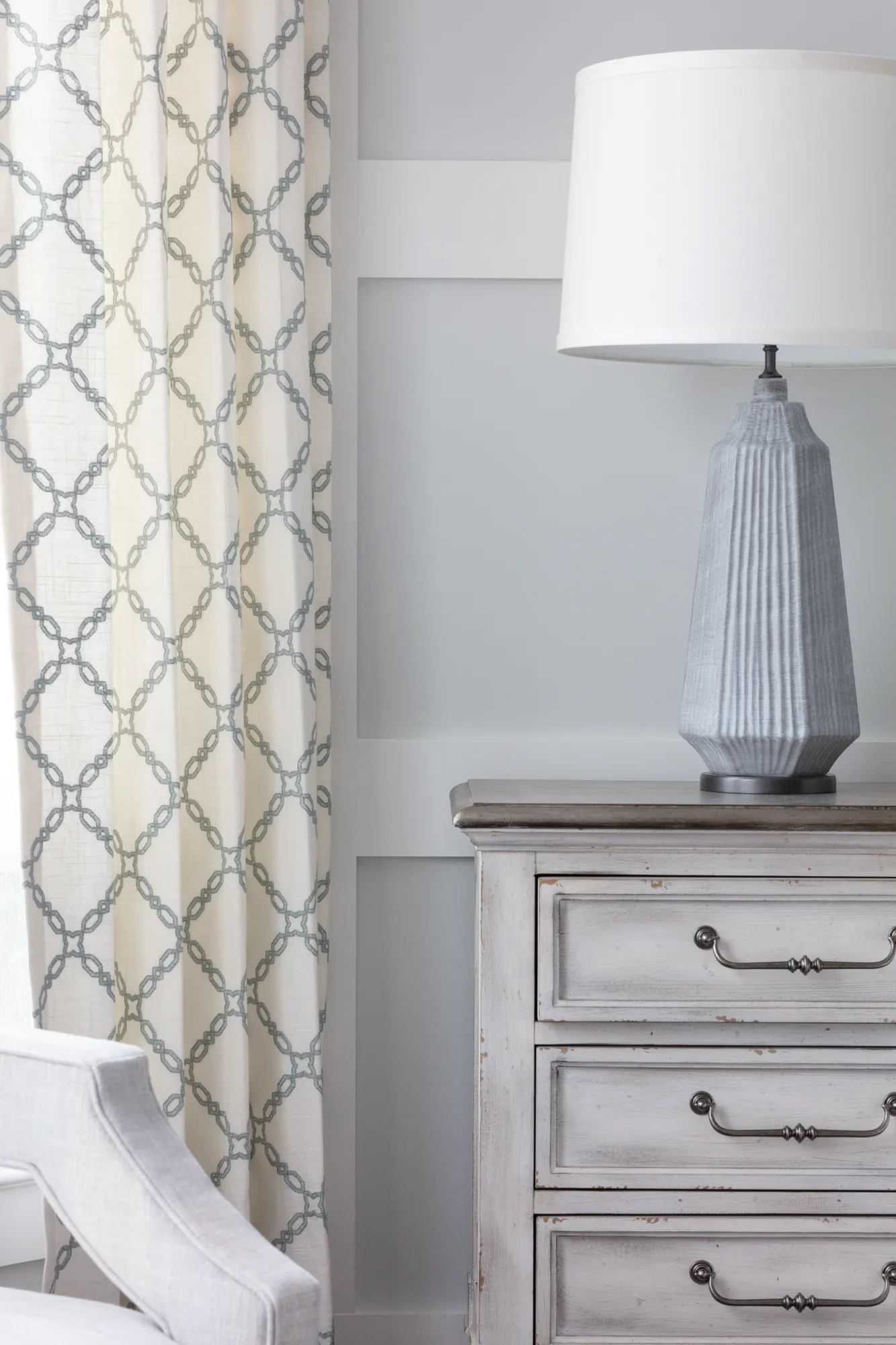 A distressed white chest of drawers holds a tall gray fluted table lamp, next to a patterned curtain and a light-colored upholstered chair.