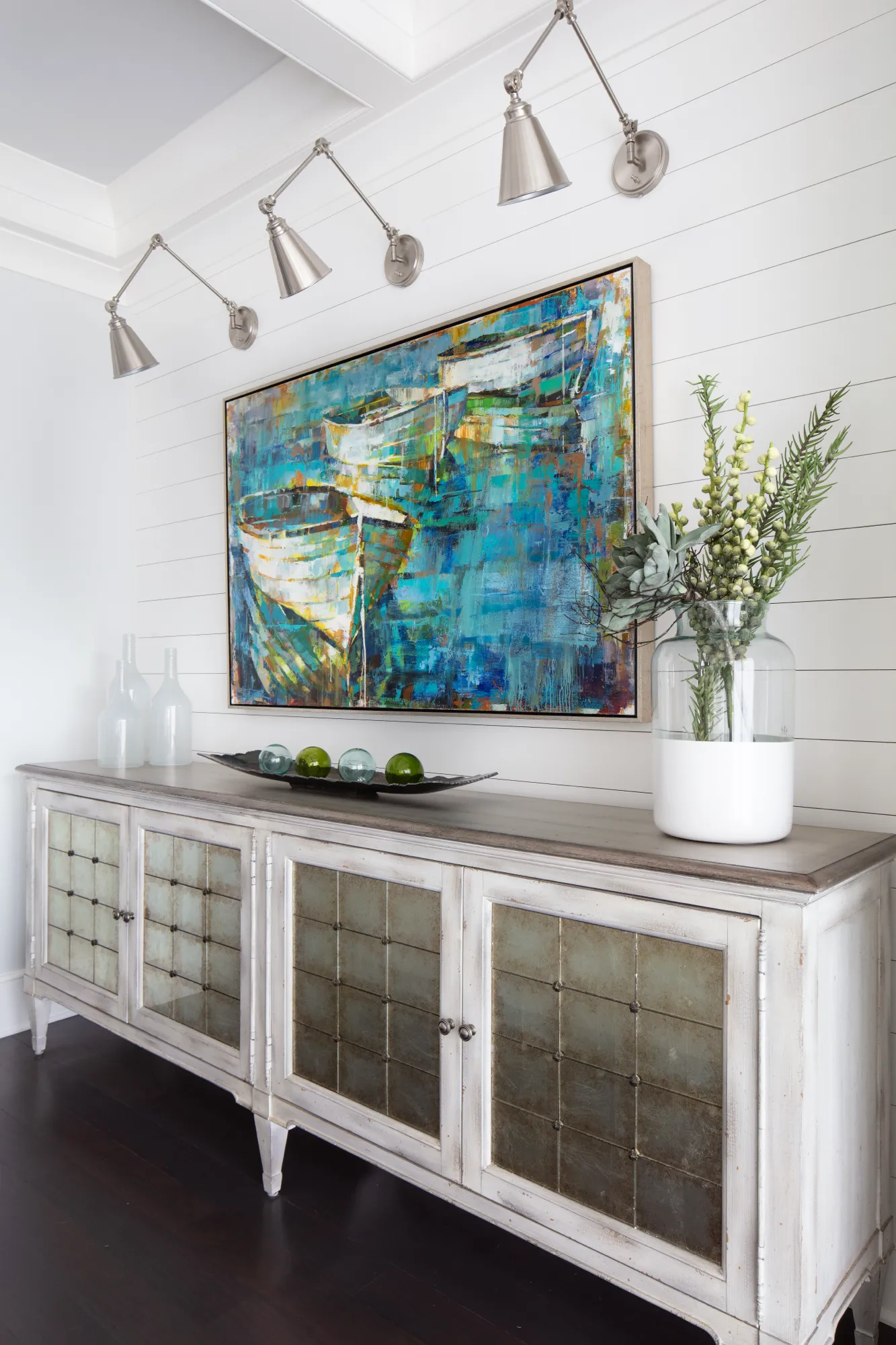 A long, distressed white credenza with mirrored doors holds a vase of greenery and decorative glass orbs, positioned beneath a vibrant abstract boat painting.