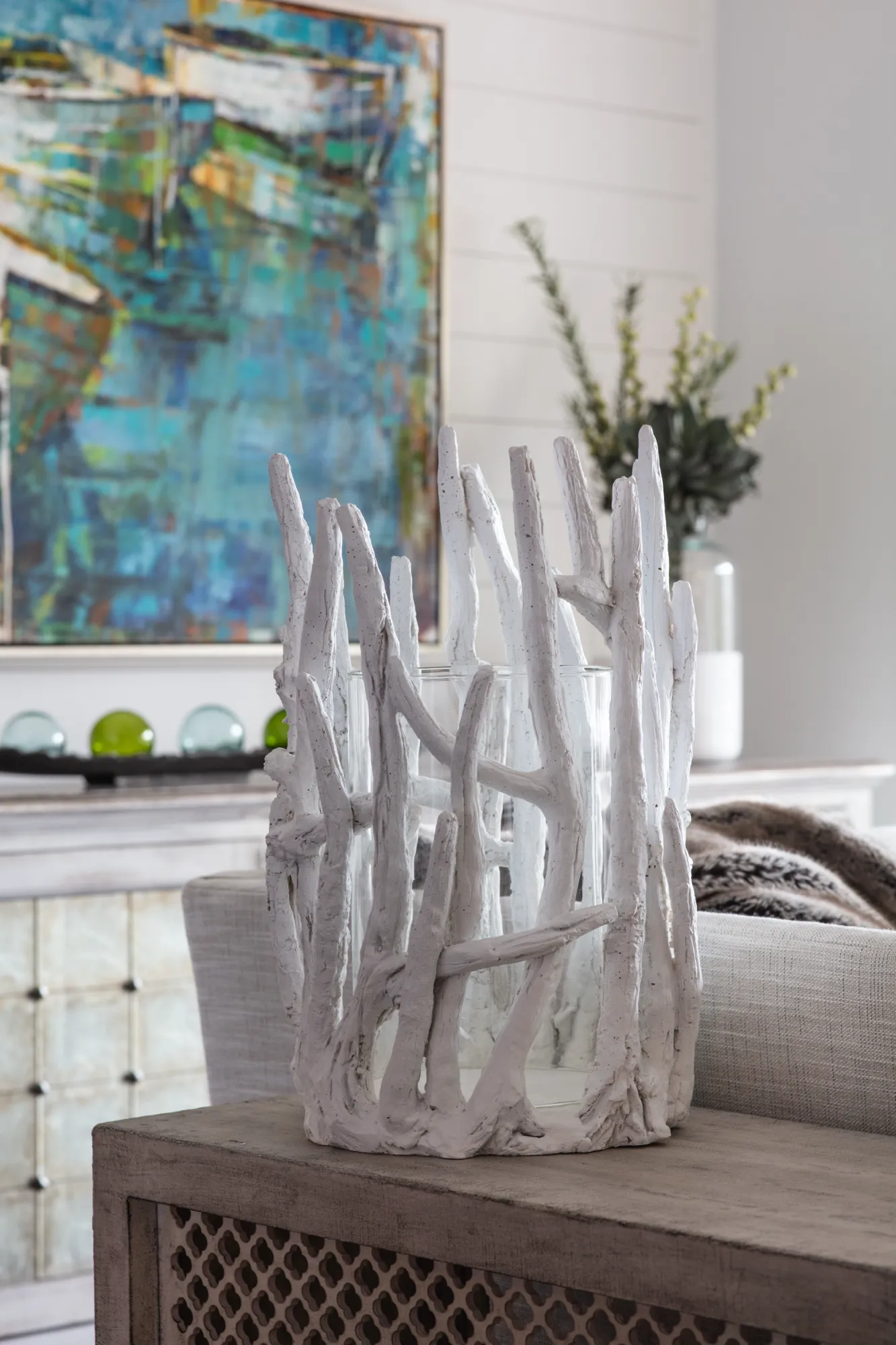 A decorative piece featuring a glass cylinder surrounded by textural white faux driftwood sits on a light gray end table. A large abstract painting is visible behind it.