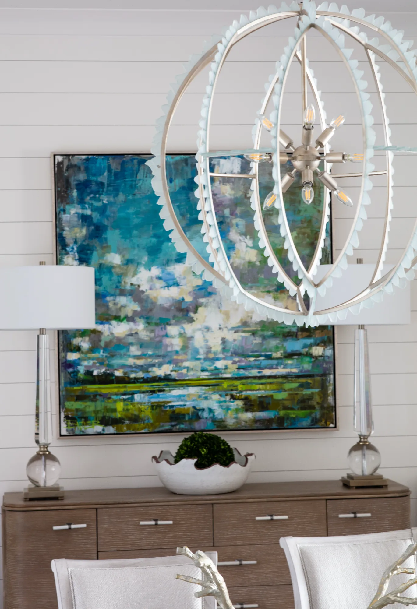 A coastal-style dining area with white shiplap walls. A unique spherical chandelier illuminates a wooden credenza displaying an abstract landscape painting and two crystal lamps.