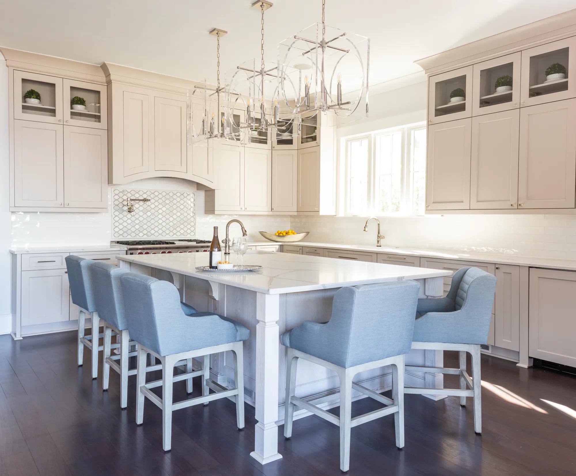 A bright, modern kitchen features light gray cabinets, a white island, and dark wood floors. Five elegant light blue upholstered bar stools sit under the island, illuminated by two clear chandeliers.