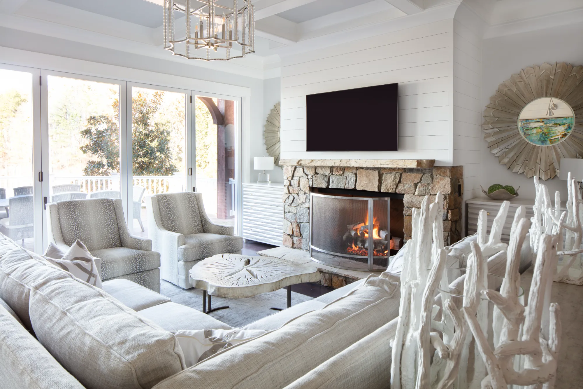 A bright, coastal-style living room with a stone fireplace, light gray sectional, and spotted accent chairs. Glass doors open onto an exterior deck.