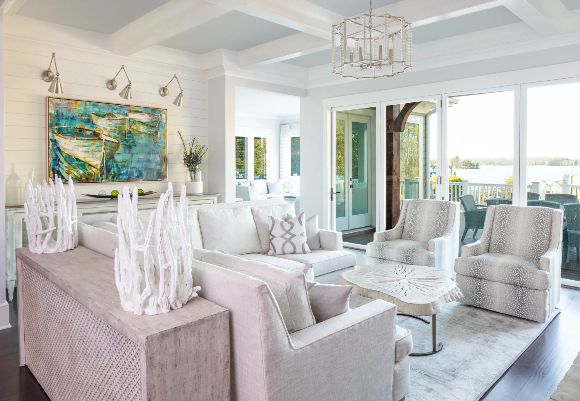 A luxurious, coastal-style living room featuring neutral furniture, white shiplap walls, and nautical art. Large glass doors open to a deck with a view of the lake.