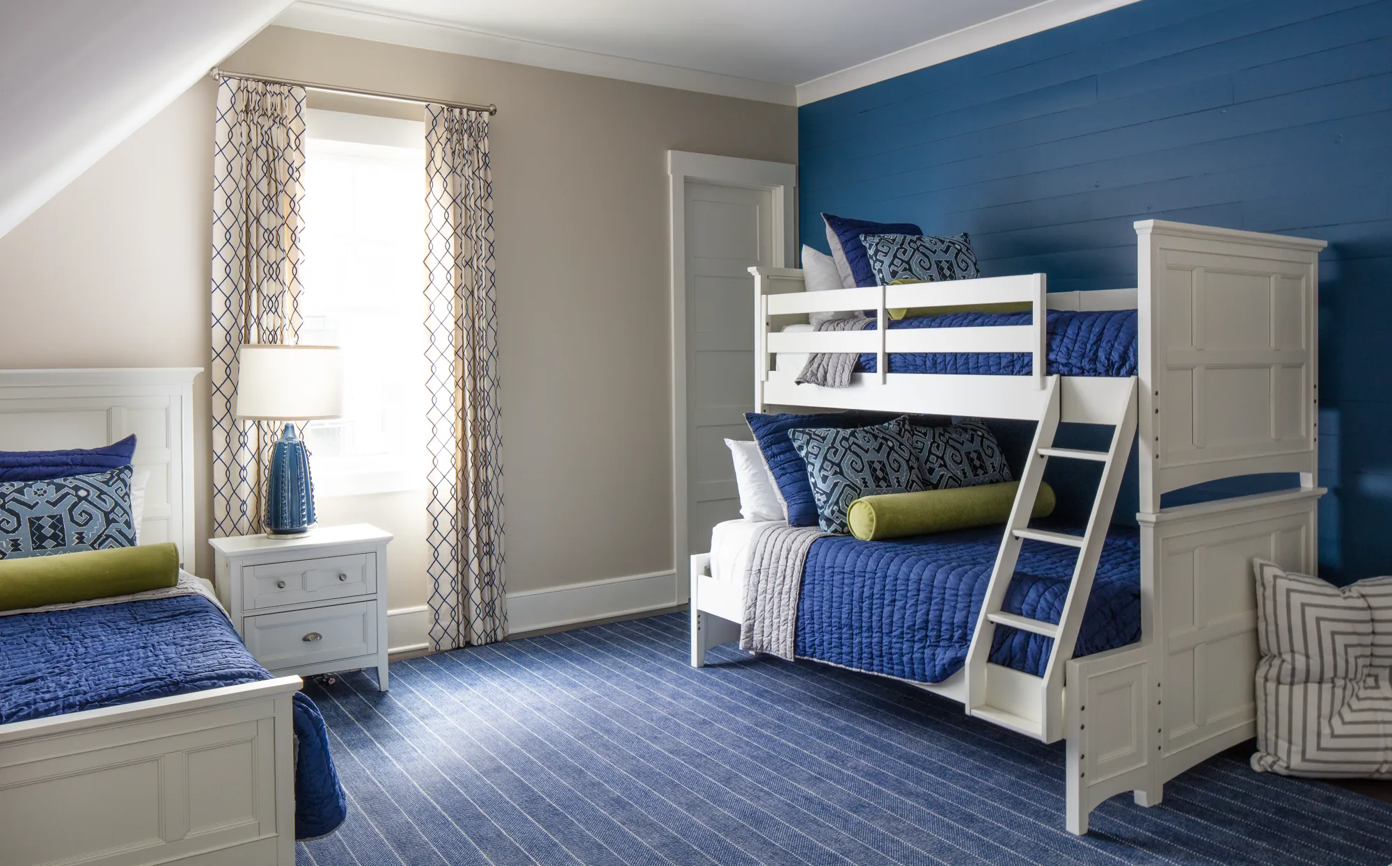 A bright, professionally designed bedroom with a white bunk bed and matching single bed. The accent wall is blue shiplap, complemented by blue bedding and a striped area rug.
