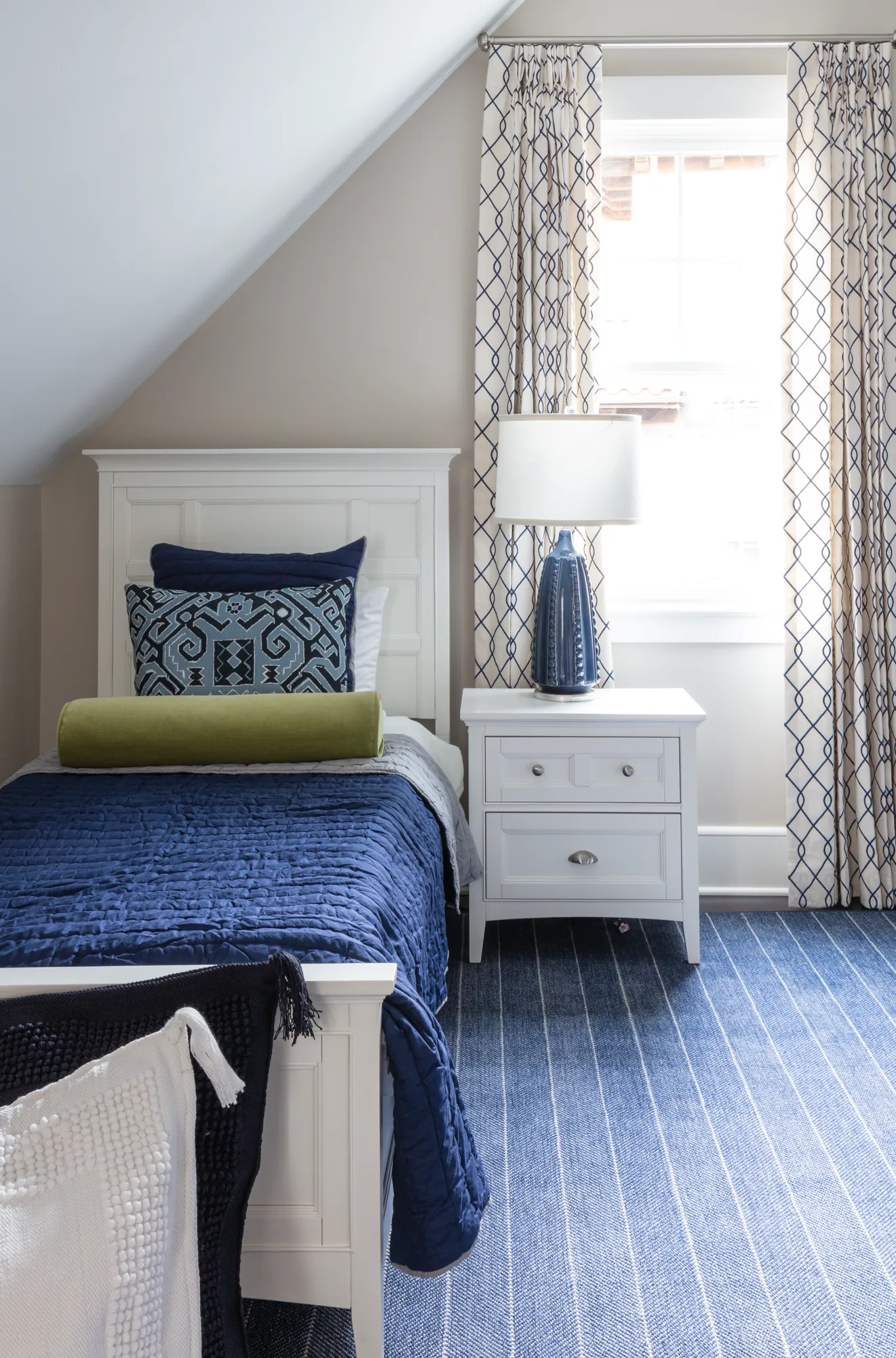 A white twin bed with a navy blue quilt and lime green bolster sits in a bedroom with a sloped ceiling. Patterned blue curtains hang next to a white nightstand holding a ceramic blue lamp.