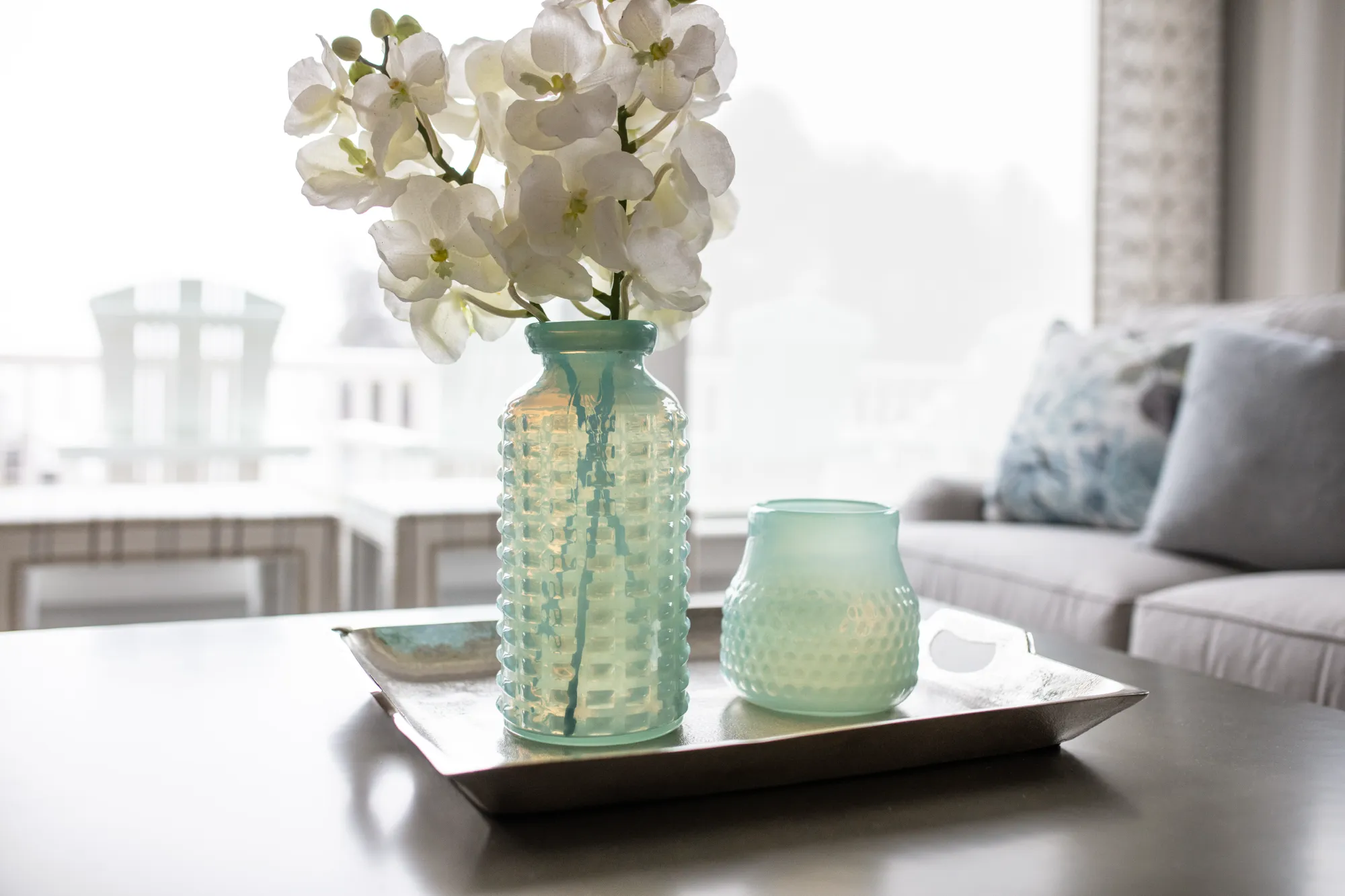 A silver tray holds two textured, aqua glass vases, one filled with white orchids, sitting on a coffee table near a light gray couch and bright window.
