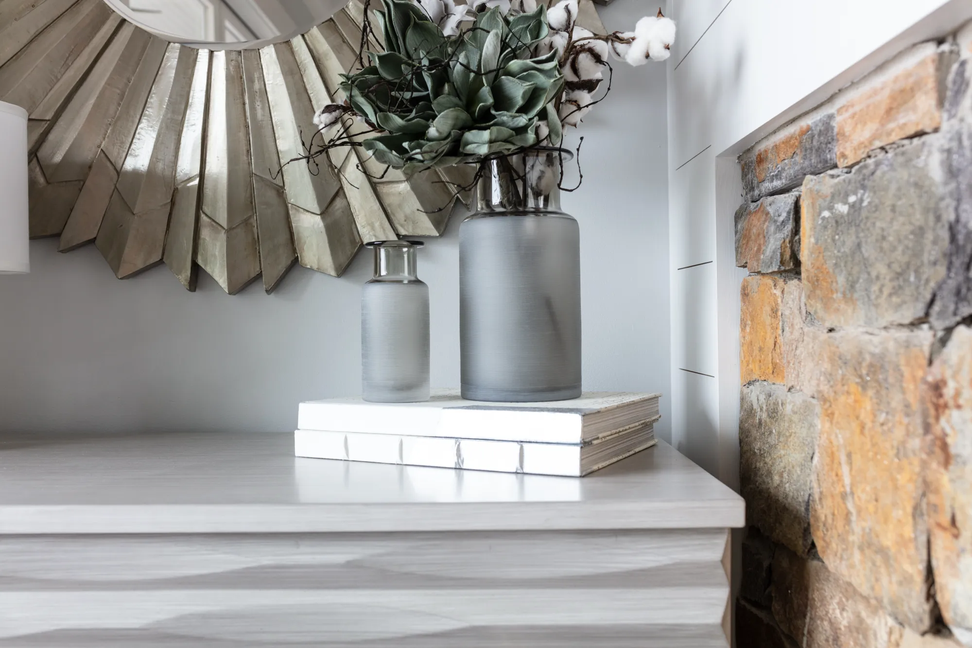 Two frosted gray vases holding succulents and cotton stems rest on white books on a light wooden surface. Behind them is a silver sunburst mirror and next to them is a rough stone fireplace surround.