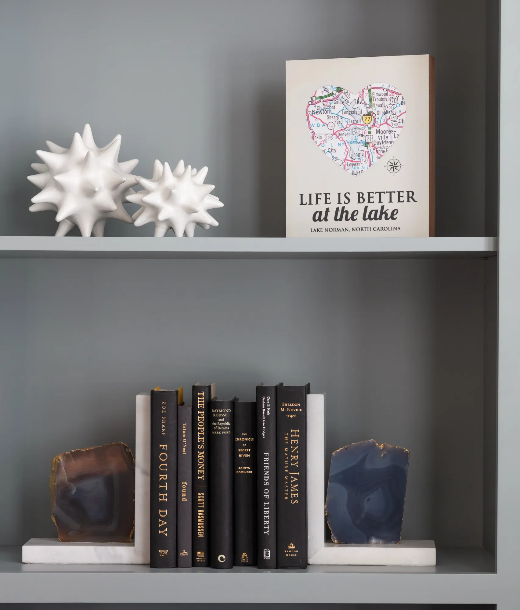 A modern gray bookshelf styled with abstract white sculptures and a Lake Norman print. Black books are secured by blue-brown agate and marble bookends.