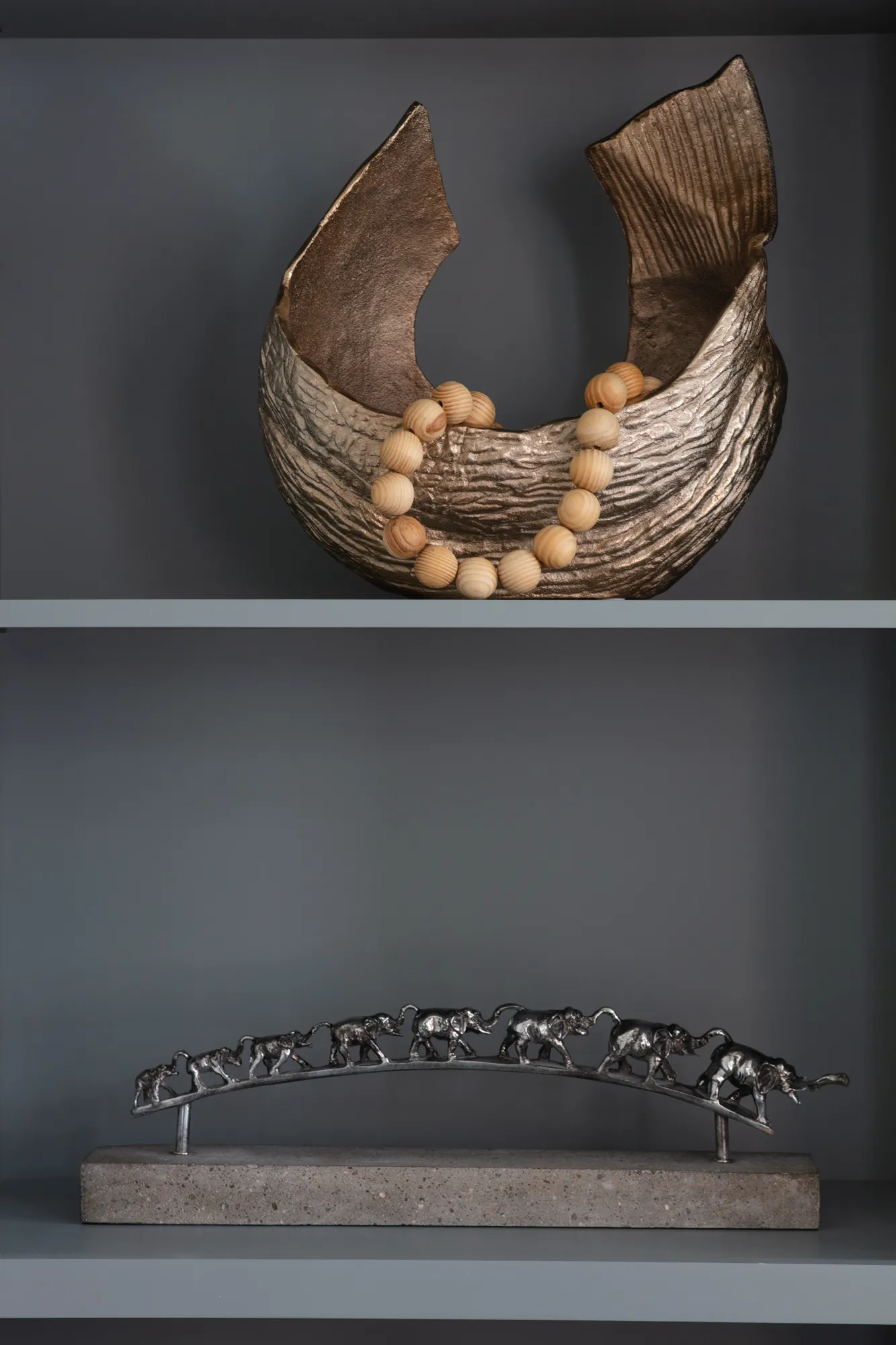 Two modern decorative objects displayed on dark gray shelving: a bronze textured bowl holding wood beads on the top, and a silver metal elephant sculpture on a stone base below.