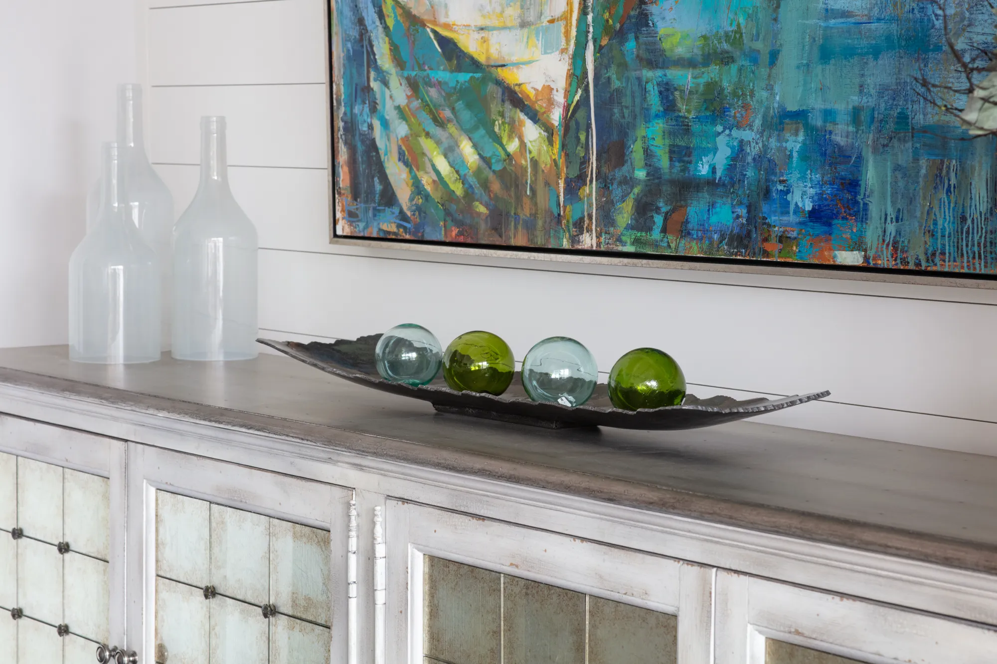 A distressed white console topped with three frosted bottles and a dish of green and blue glass spheres, set beneath an abstract painting on a shiplap wall.