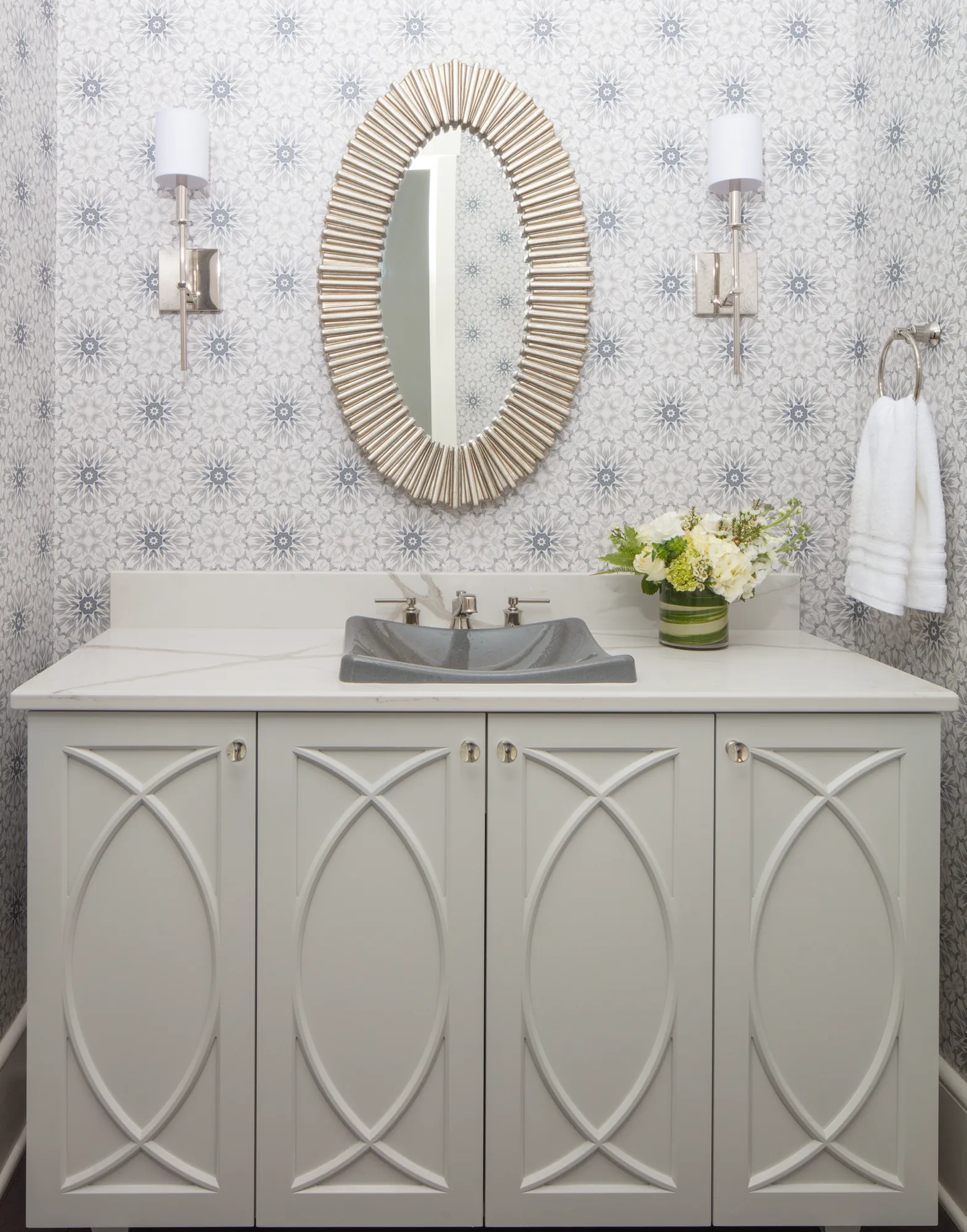A white vanity with geometric molding and a gray contoured sink sits beneath an oval mirror. The walls feature light gray patterned wallpaper.