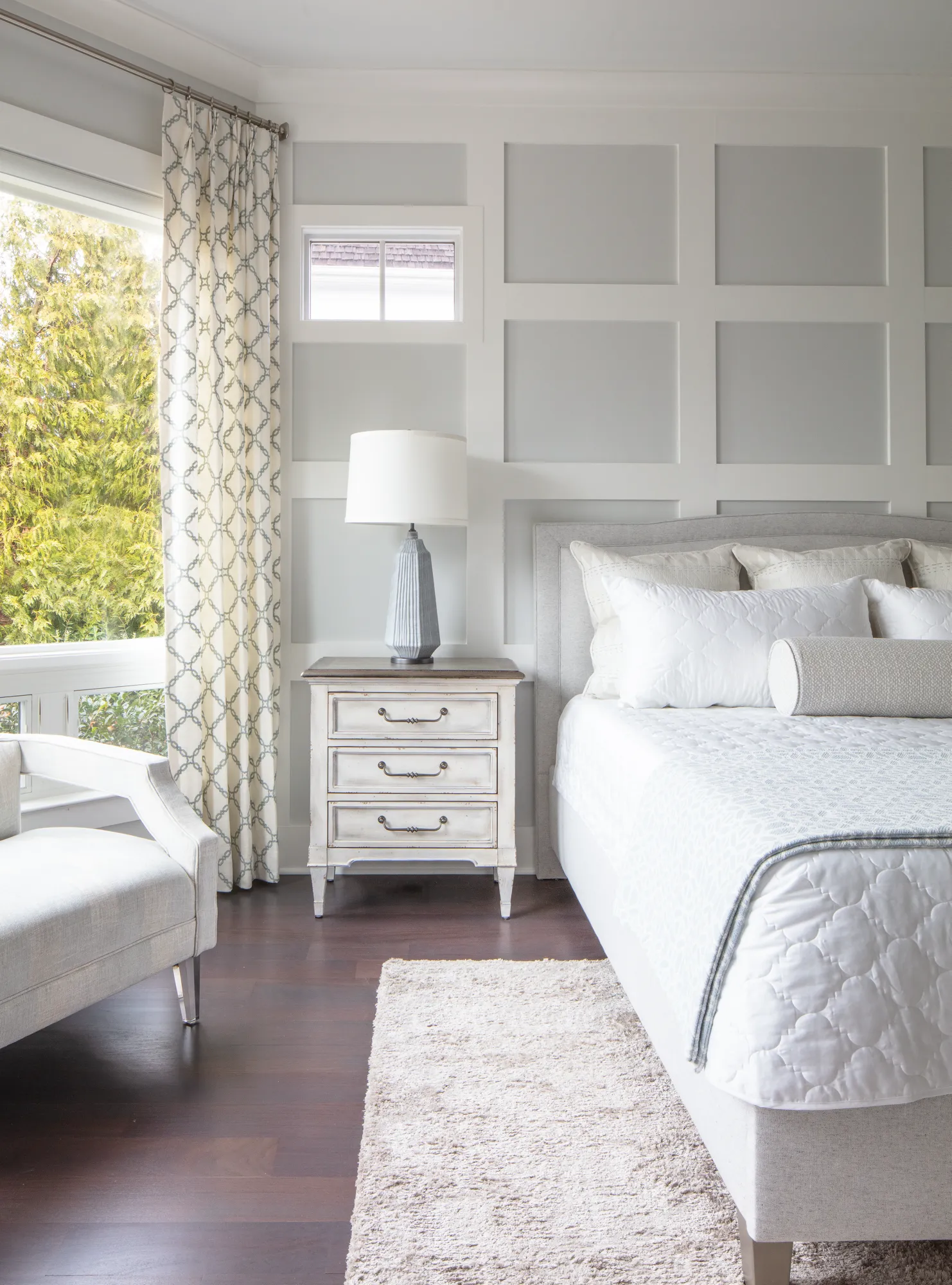 A bright bedroom with dark wood floors and a plush white bed. A wainscoted wall acts as a headboard backdrop. A large window with patterned curtains provides natural light.