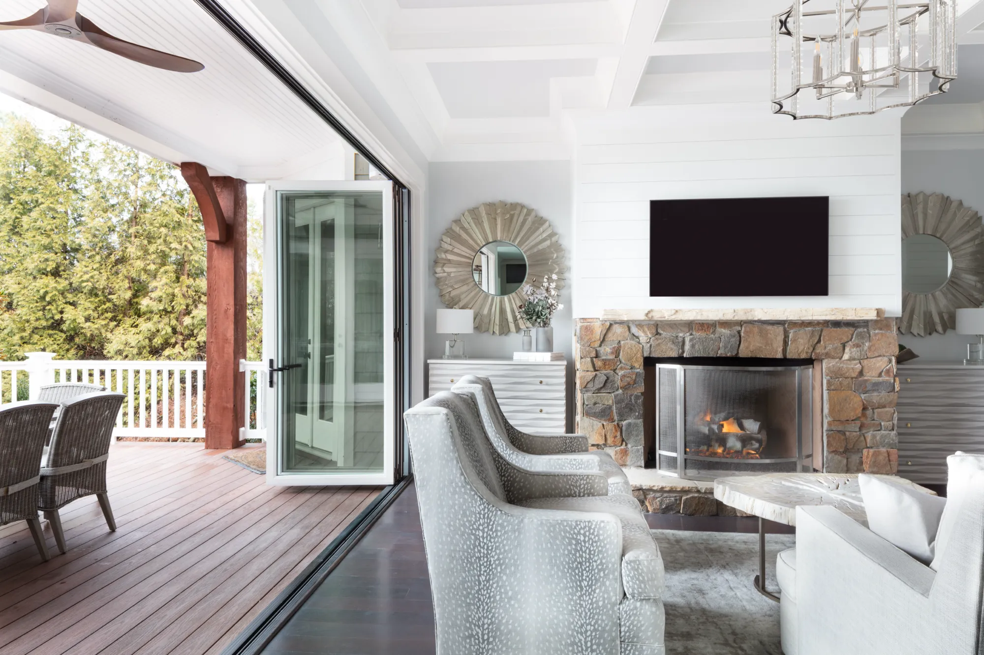An elegant living room opens fully to a wooden deck and wooded view. The interior features white shiplap walls, a lit stone fireplace, and patterned high-backed chairs.