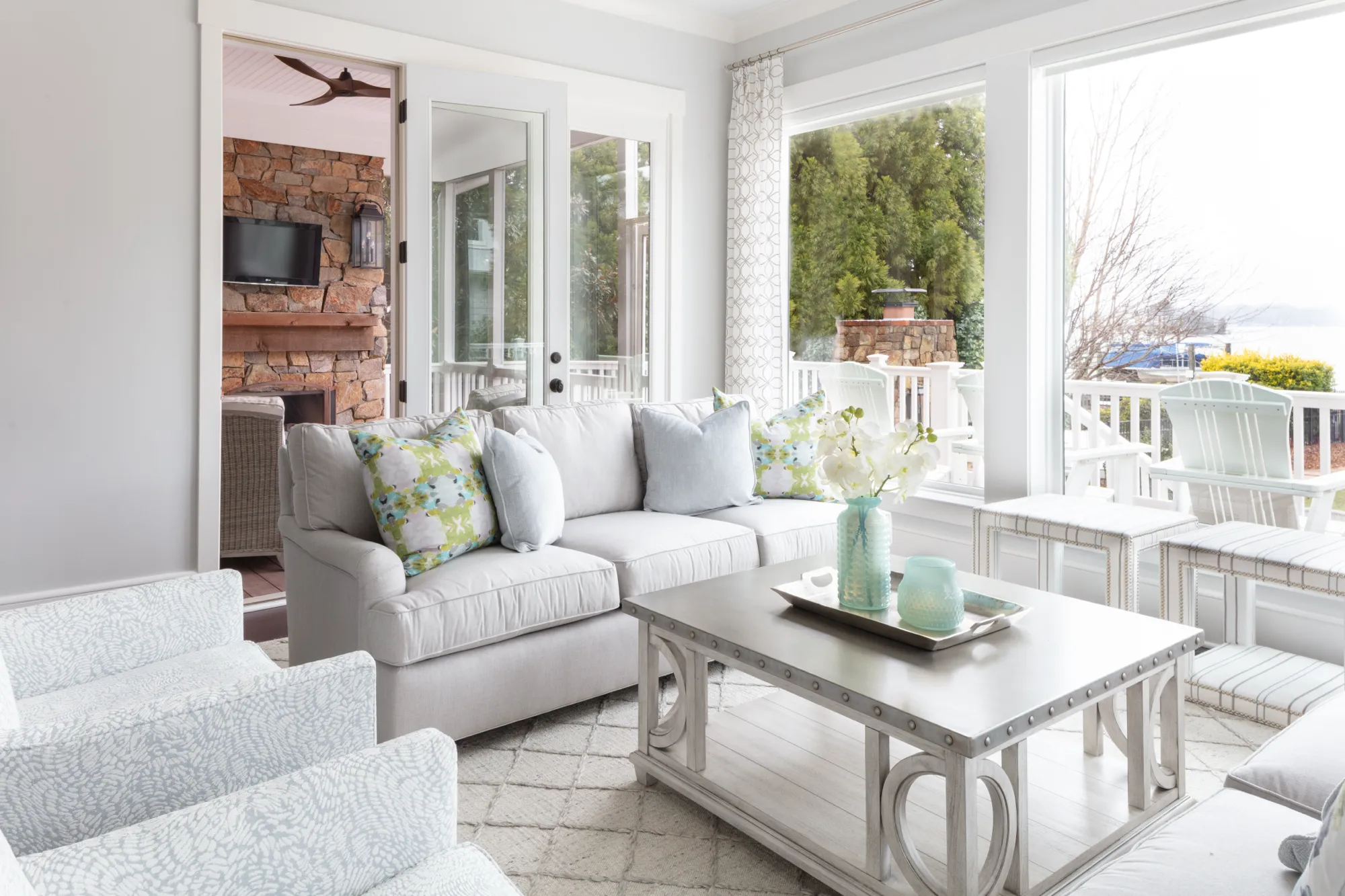 A bright, airy sunroom featuring a neutral sofa with green and blue accent pillows, a decorative coffee table, and large windows offering a view of a deck and lake.