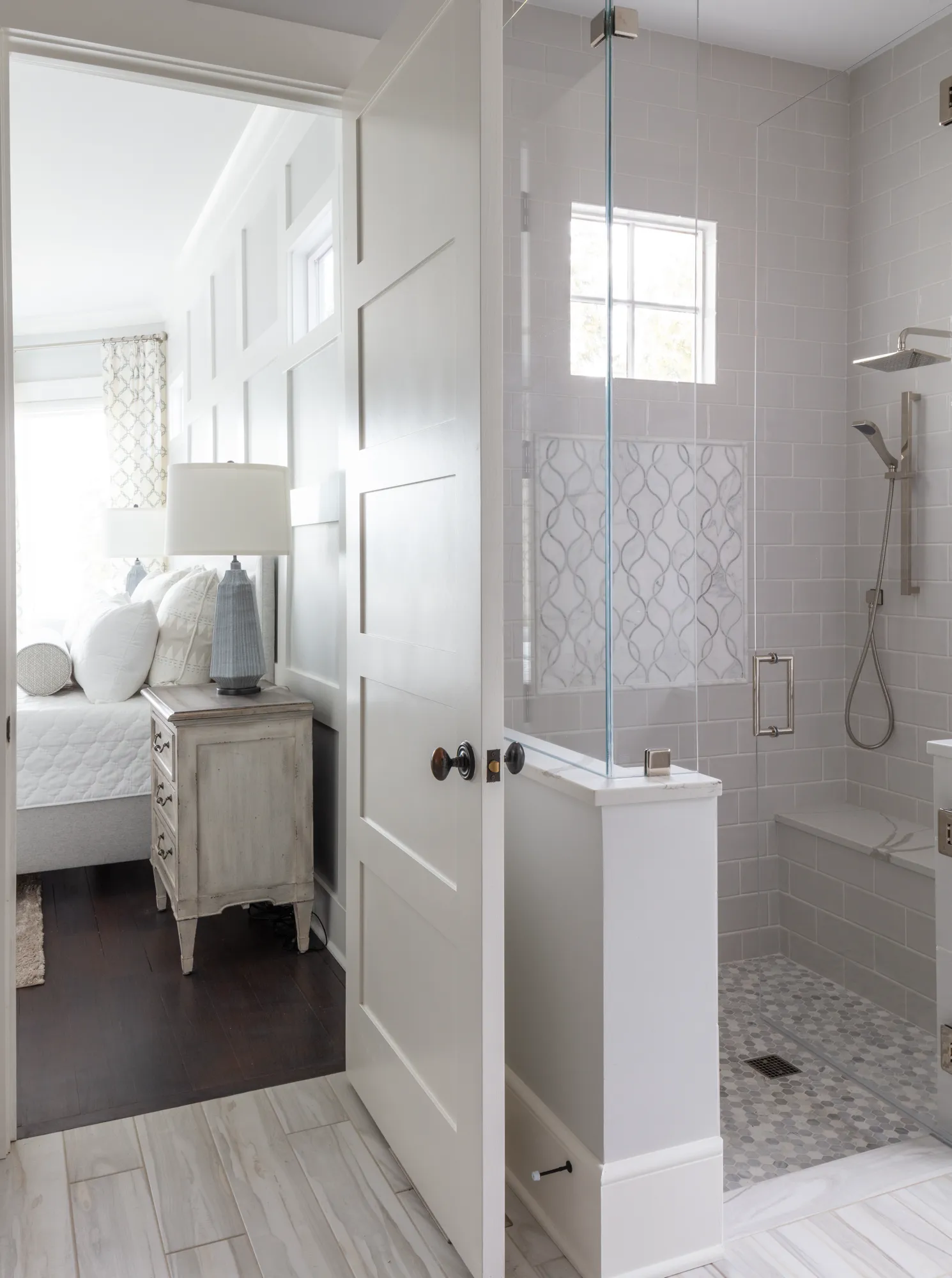 A modern bathroom features gray subway tile and a glass walk-in shower. An open white door reveals a bright bedroom with a bed, white linens, and a distressed nightstand.