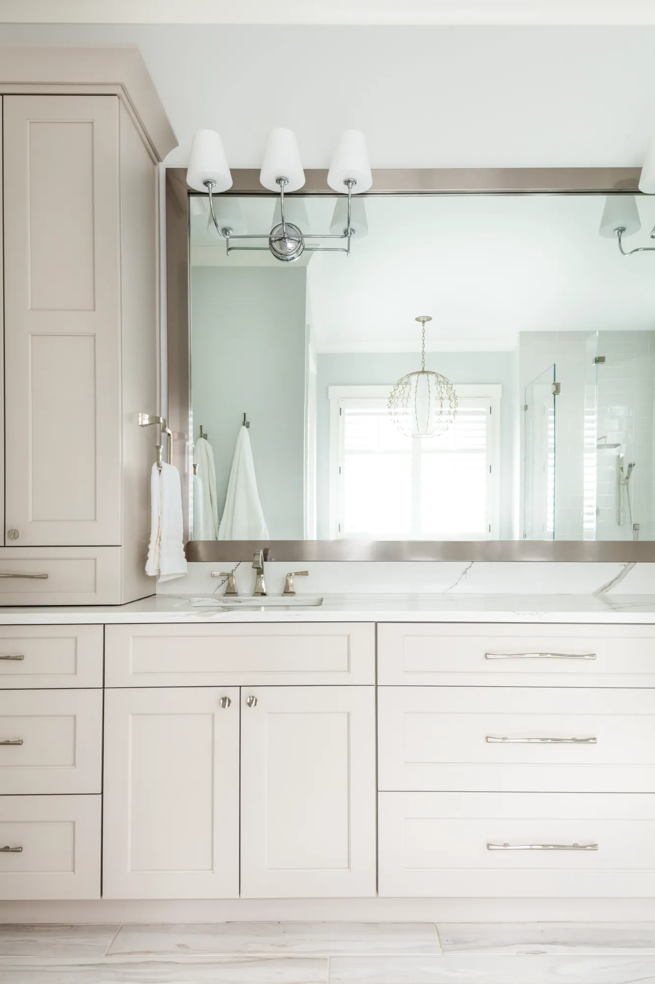 A neutral modern bathroom vanity with greige cabinetry, a white marble counter, and a large mirror reflecting a window and a sparkling chandelier.