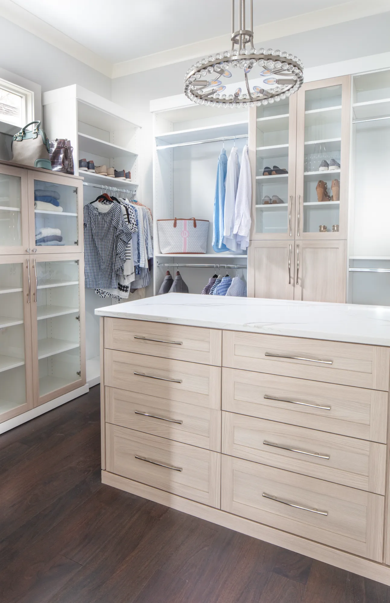 A spacious, custom walk-in closet featuring light wood cabinetry, a large central island with a white top, and dark wood flooring. Clothes are neatly organized.