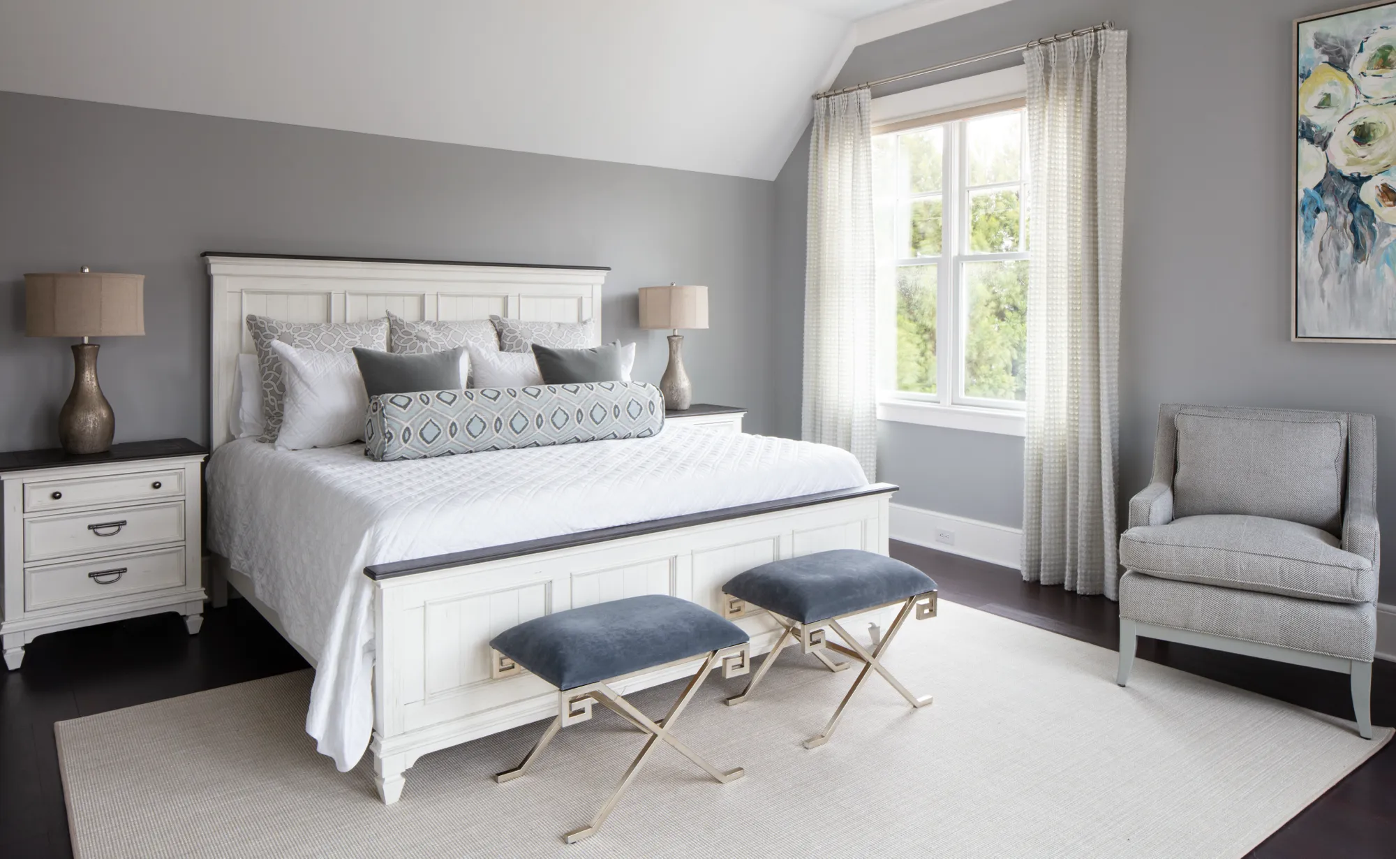A white bed with gray pillows and blue velvet stools sits centered in a serene bedroom with light gray walls. Natural light floods in from a window next to a gray upholstered chair.