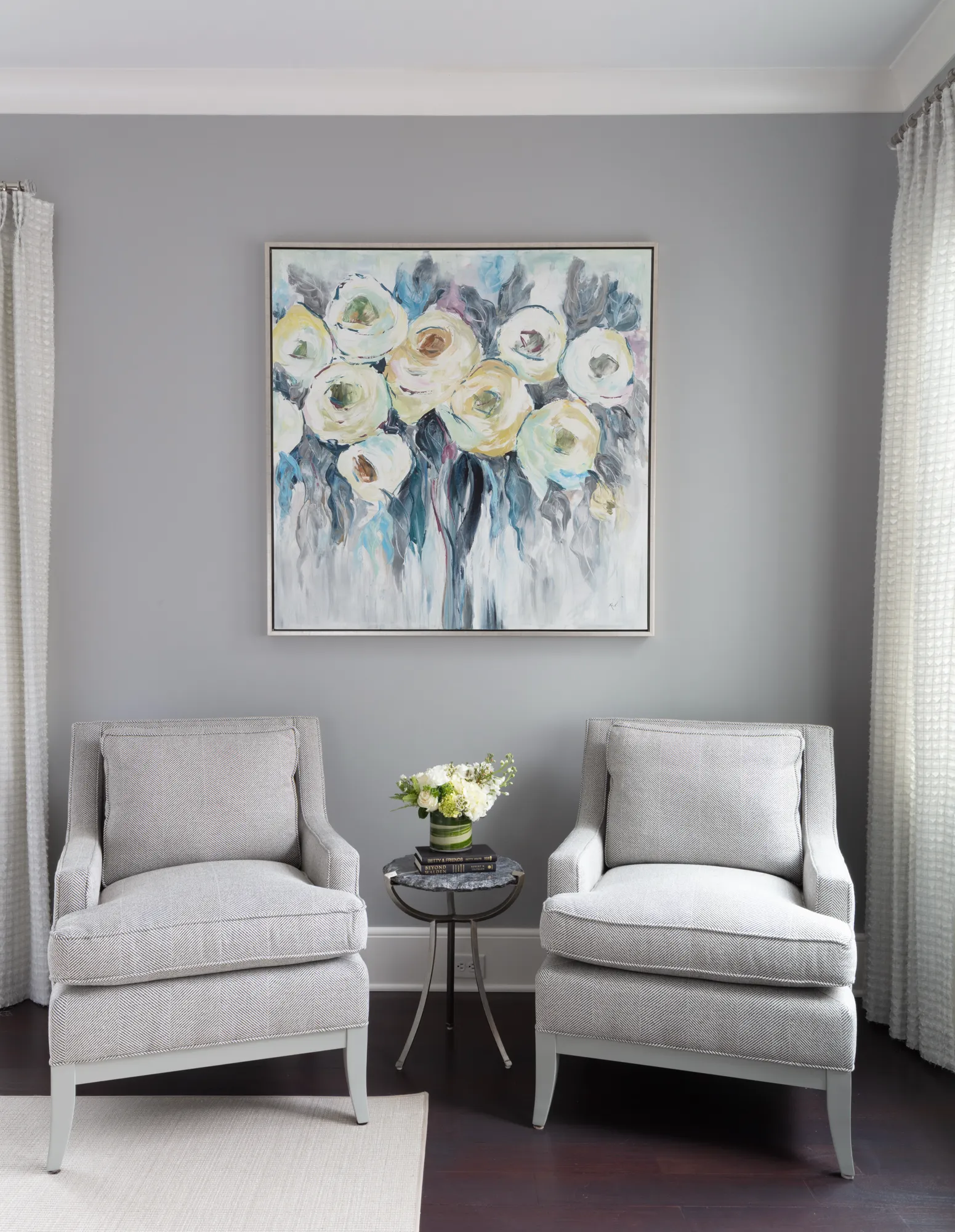 A seating nook with two herringbone gray armchairs, separated by a side table with flowers, centered below a large abstract floral painting.