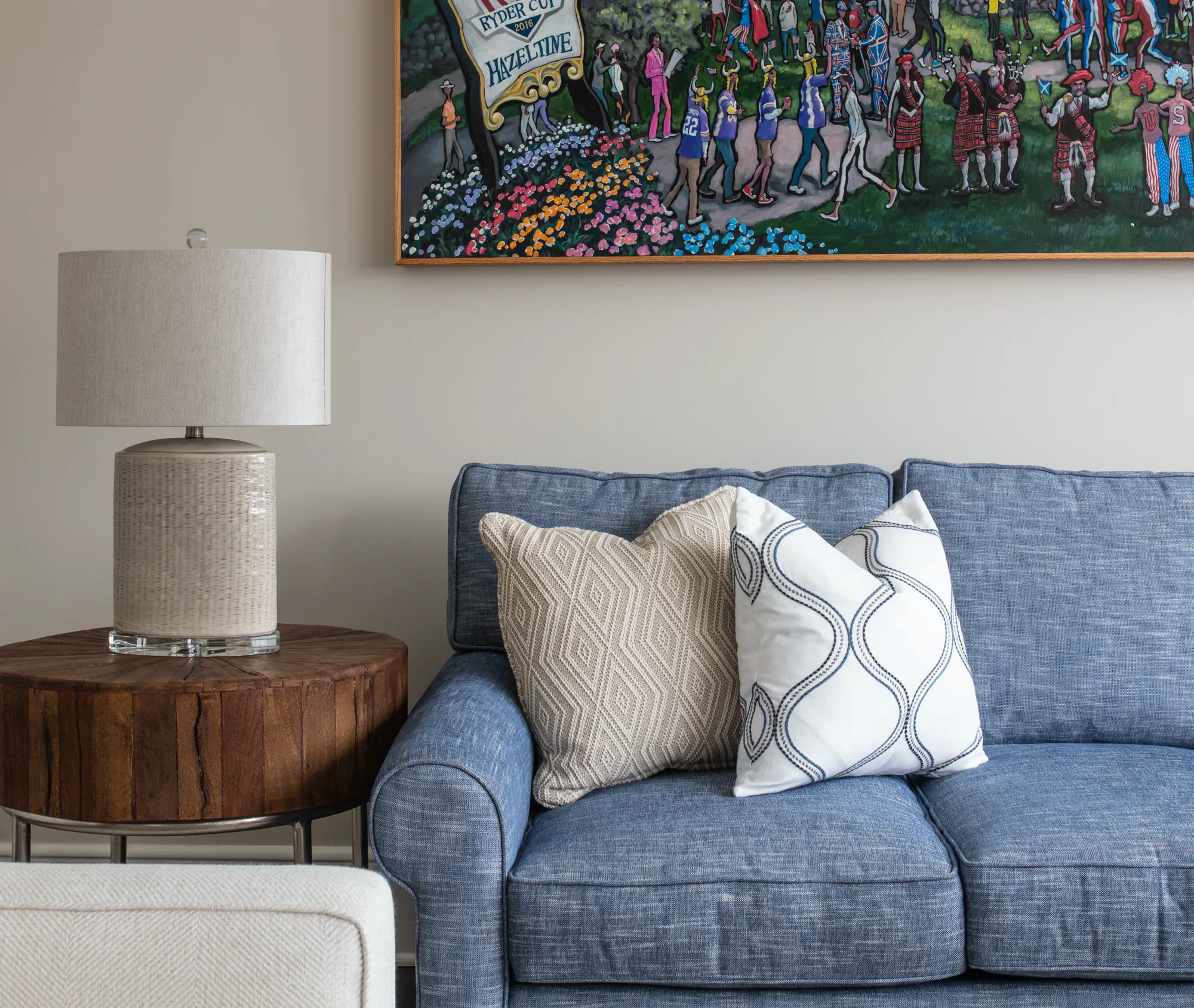 A comfortable blue sofa is dressed with two patterned pillows. Next to it, a round, wood side table holds a textured ceramic lamp, below a colorful golf-themed painting.