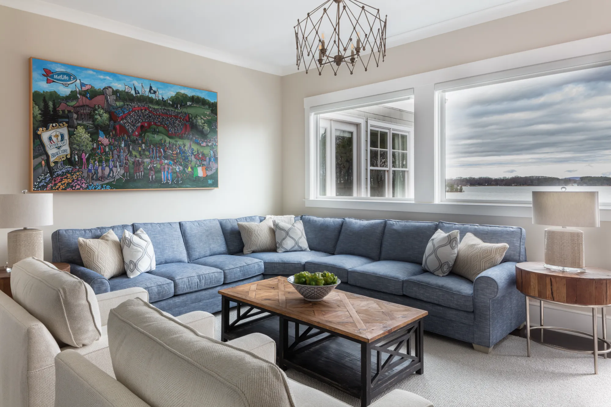 An elegant living room with a spacious blue sectional sofa, a wooden parquet coffee table, and a large decorative golf tournament painting. Lake views are visible through the windows.
