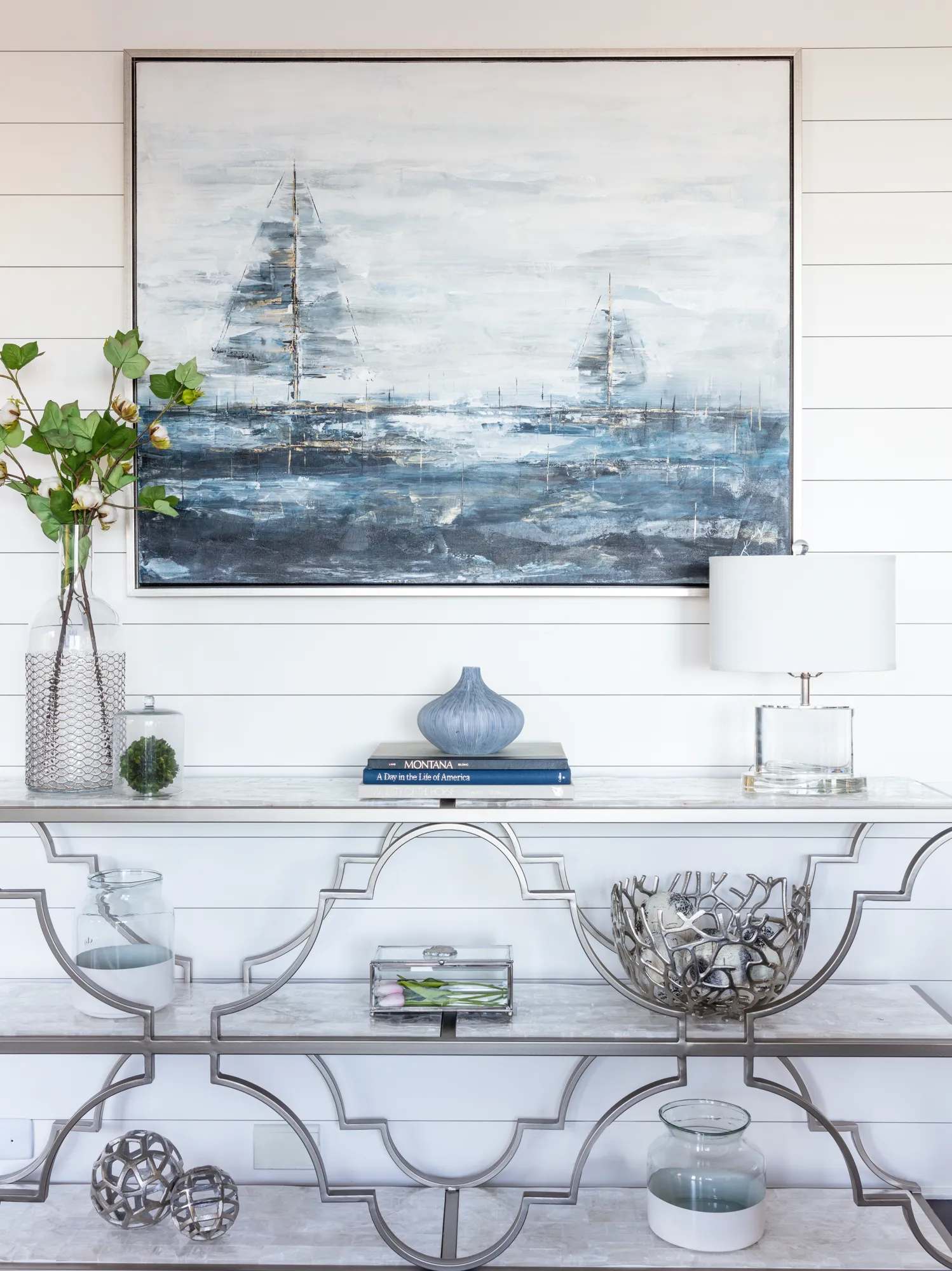 A silver decorative console table sits below an abstract painting of sailboats. The table holds a clear lamp, blue vase, books, and green branches in a glass vase, all against a white shiplap wall.