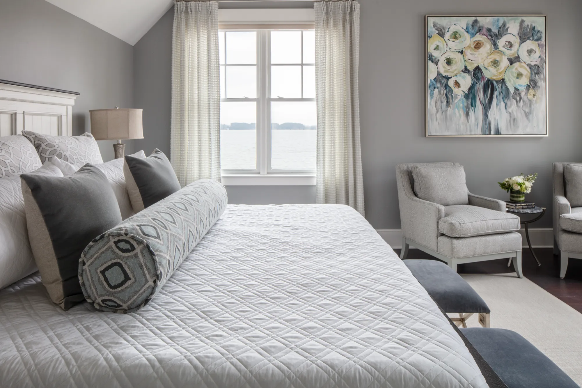 A luxurious gray and white bedroom interior. The bed is covered in a white quilted spread and features layered pillows. A large window overlooks a lake; an armchair and floral art complete the space.