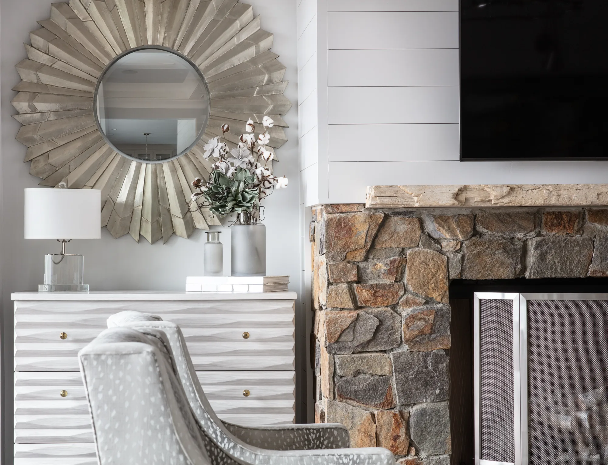 A contemporary living space featuring a large sunburst mirror above a textured white dresser, situated next to a natural stone fireplace.