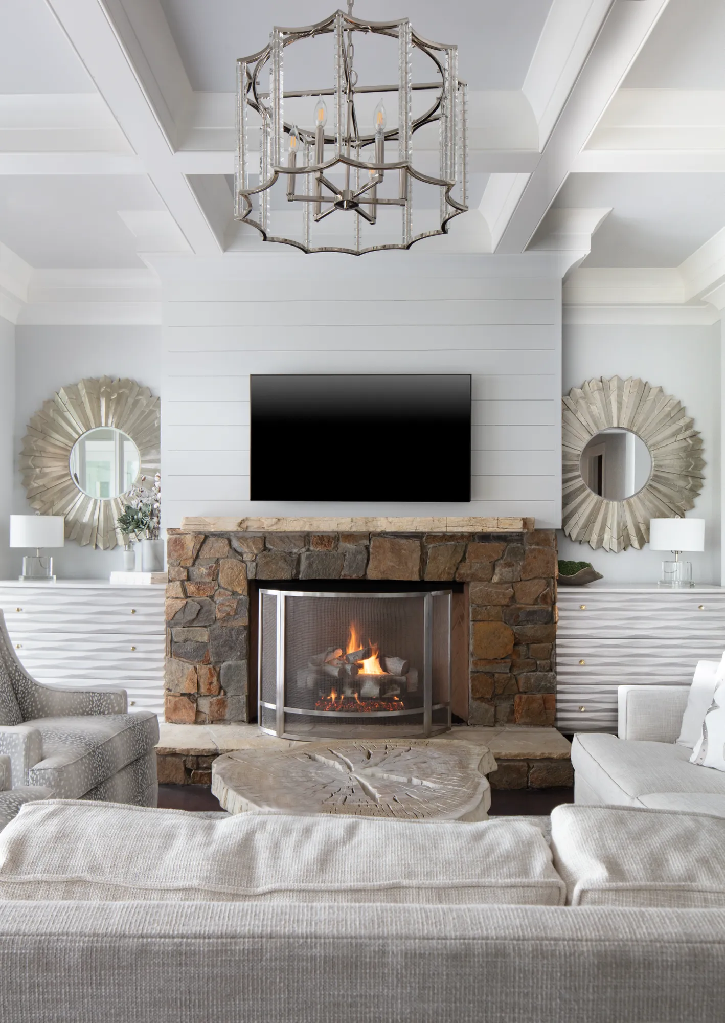 A symmetrical living room features a roaring stone fireplace with a mounted television above shiplap paneling. Sunburst mirrors flank the hearth.