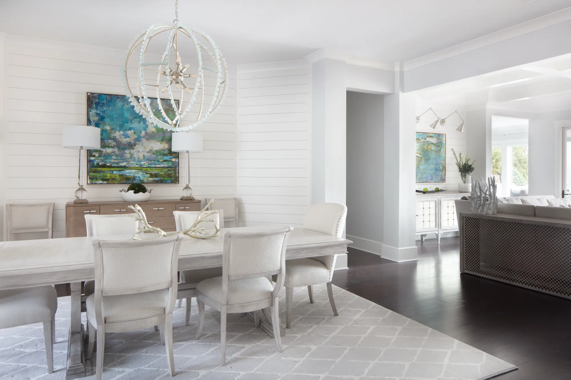 A light and elegant dining room with a gray wooden table and white upholstered chairs. A spherical chandelier and a blue abstract painting accent the white shiplap walls.