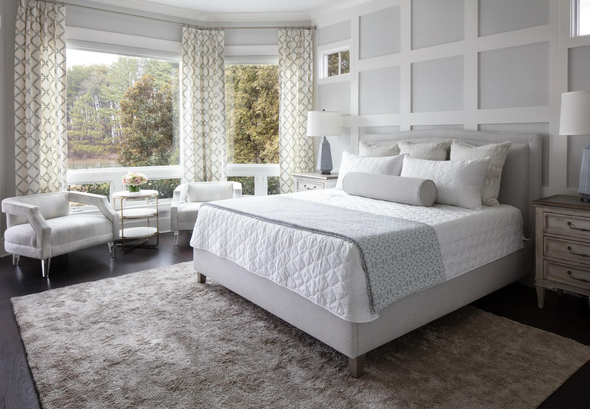 A bright, luxurious bedroom featuring a bed with white bedding and light gray accents. Large bay windows overlook a wooded landscape. The room has light gray paneled walls and a matching rug.