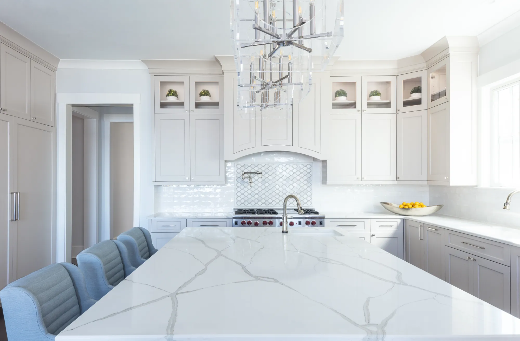 A modern kitchen featuring light gray cabinets, white marble countertops, and an island with blue upholstered seating, centered under clear acrylic chandeliers.