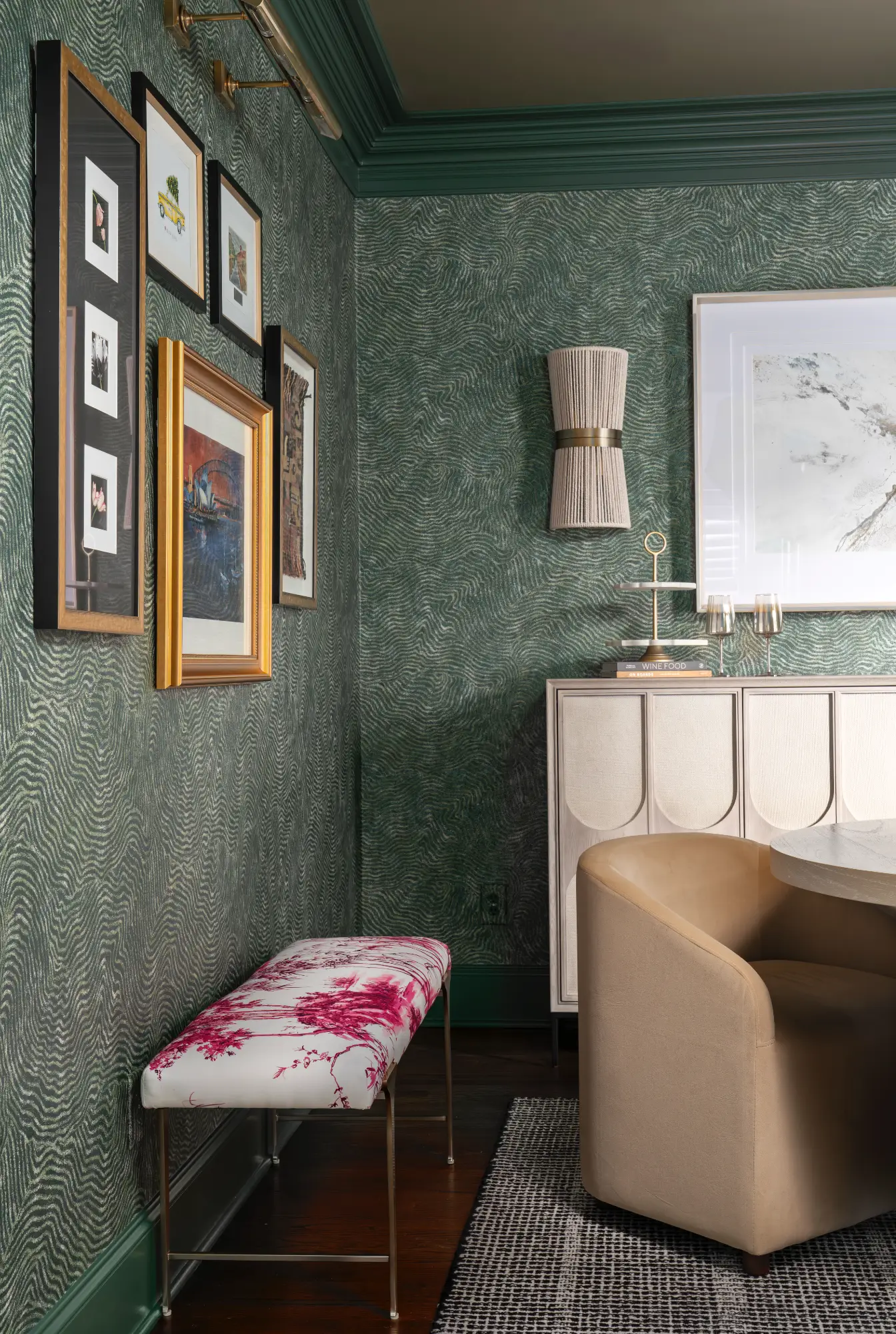 A rich, elegant interior space with green patterned wallpaper and matching molding. A gallery wall hangs over a pink floral bench, across from a beige sideboard and tan upholstered chair.